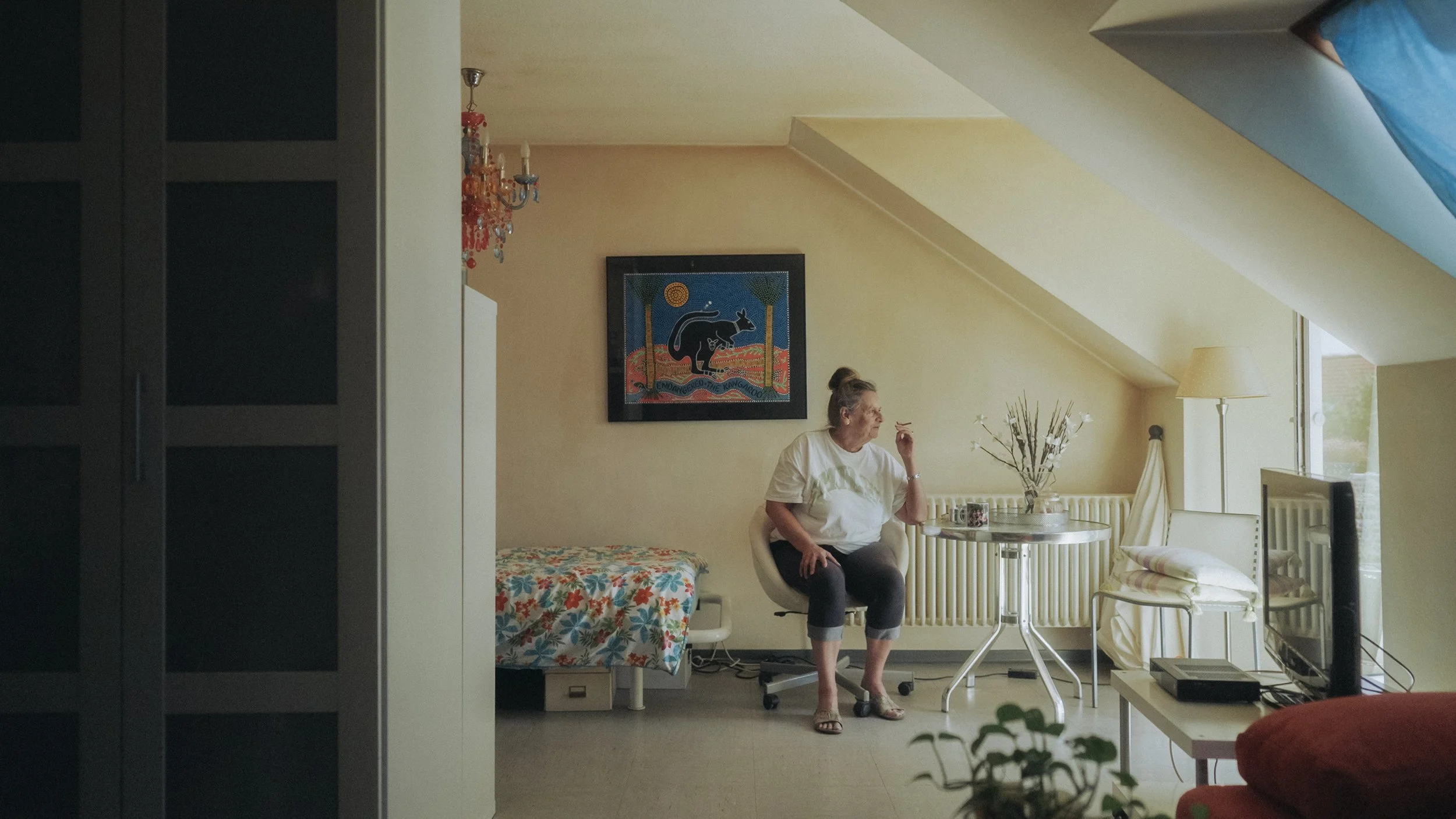 Ältere Frau sitzt in einem Sessel im Zimmer, neben einem Tisch mit blühenden Zweigen, vor einem Fenster. Es gibt ein Bett mit buntem Bezug, einen Fernseher und eine Wanddekoration mit einem Känguru-Bild.
