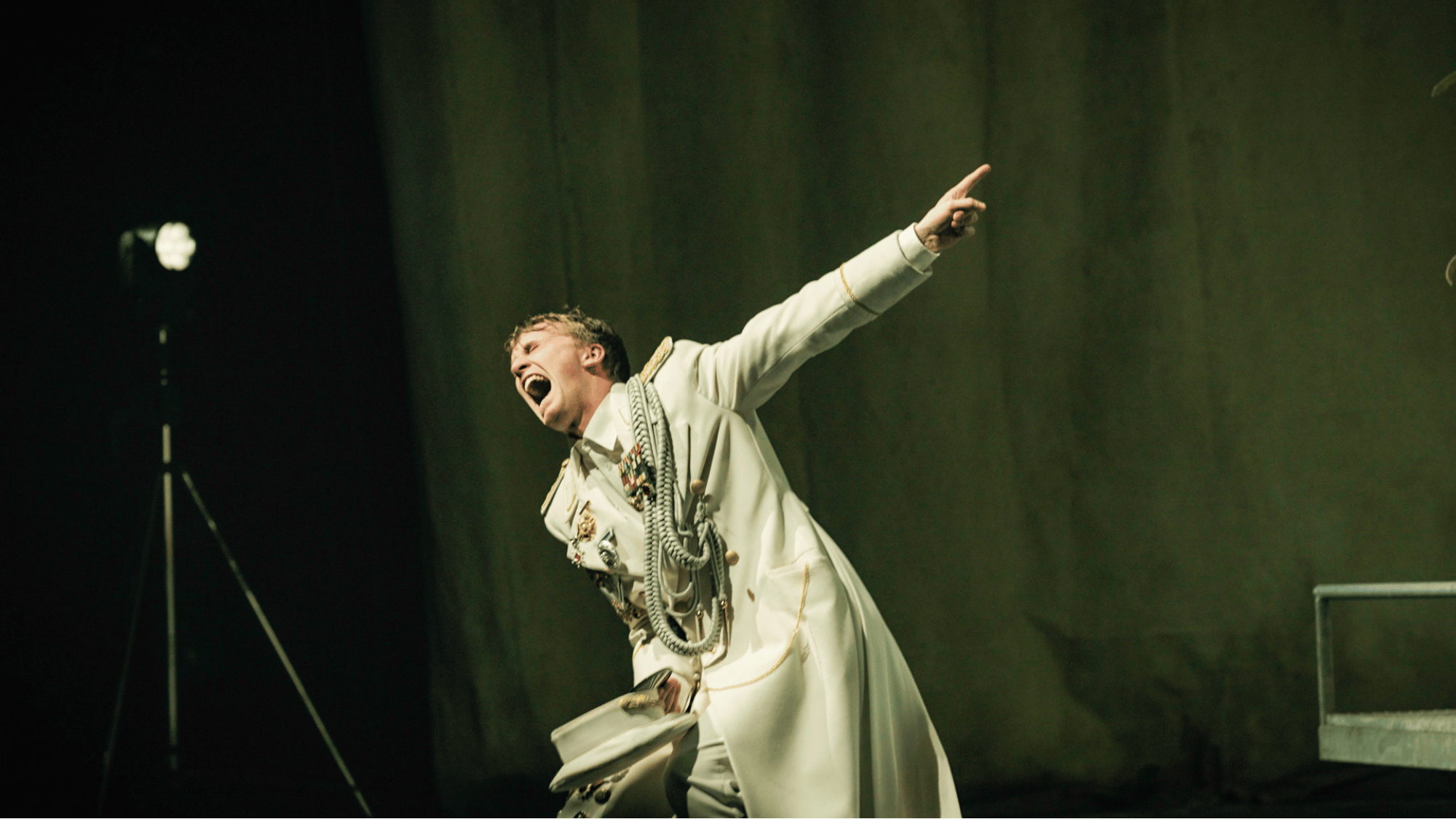 Berliner Ensemble. Ein Mann trägt ein weißes Kostüm mit Accessoires und schreit leidenschaftlich auf einer Bühne.