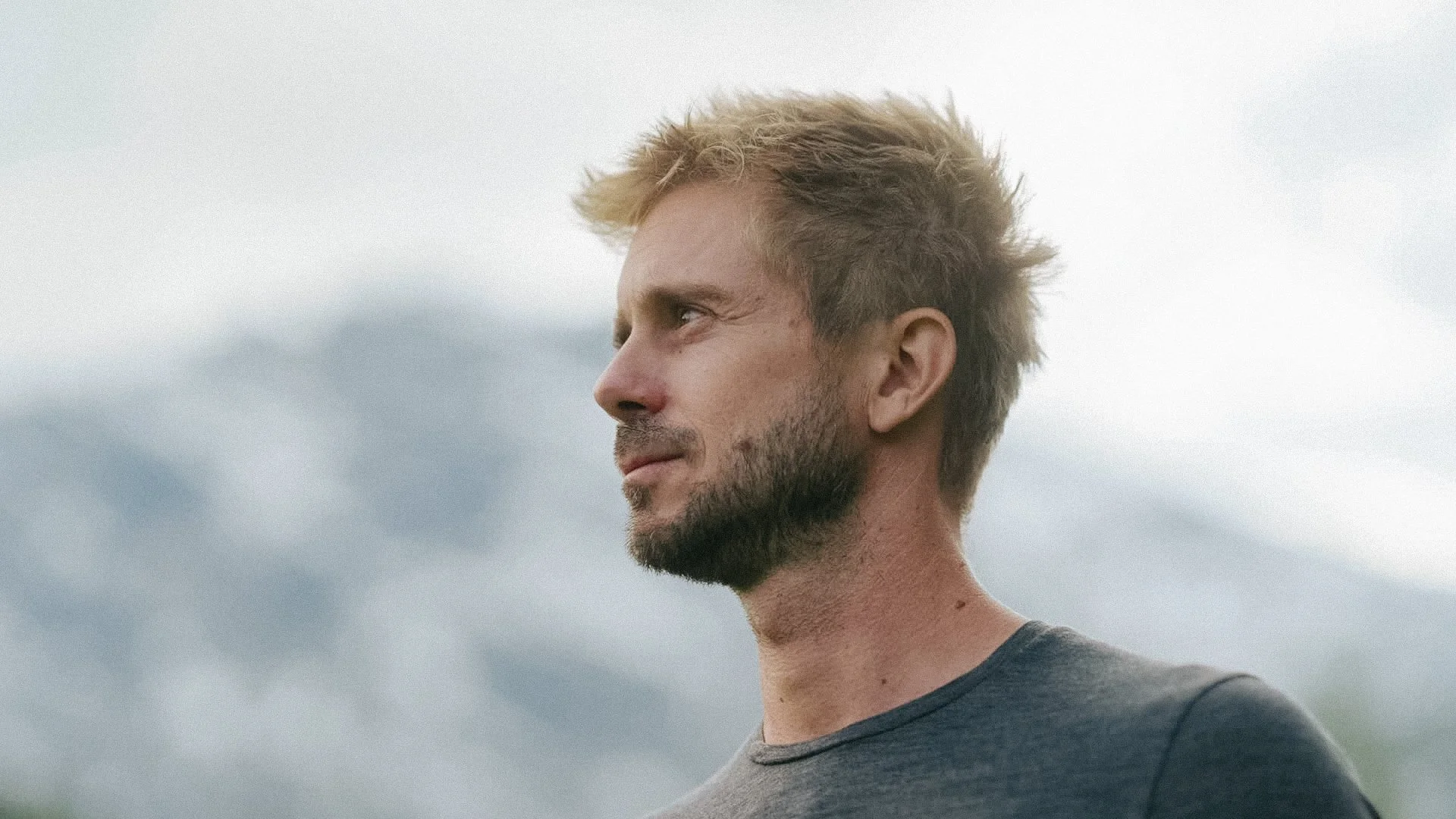 Seitenansicht eines Mannes mit blondem, etwas zerzaustem Haar, Bart und dunklem T-Shirt vor einem bewölkten Himmel.