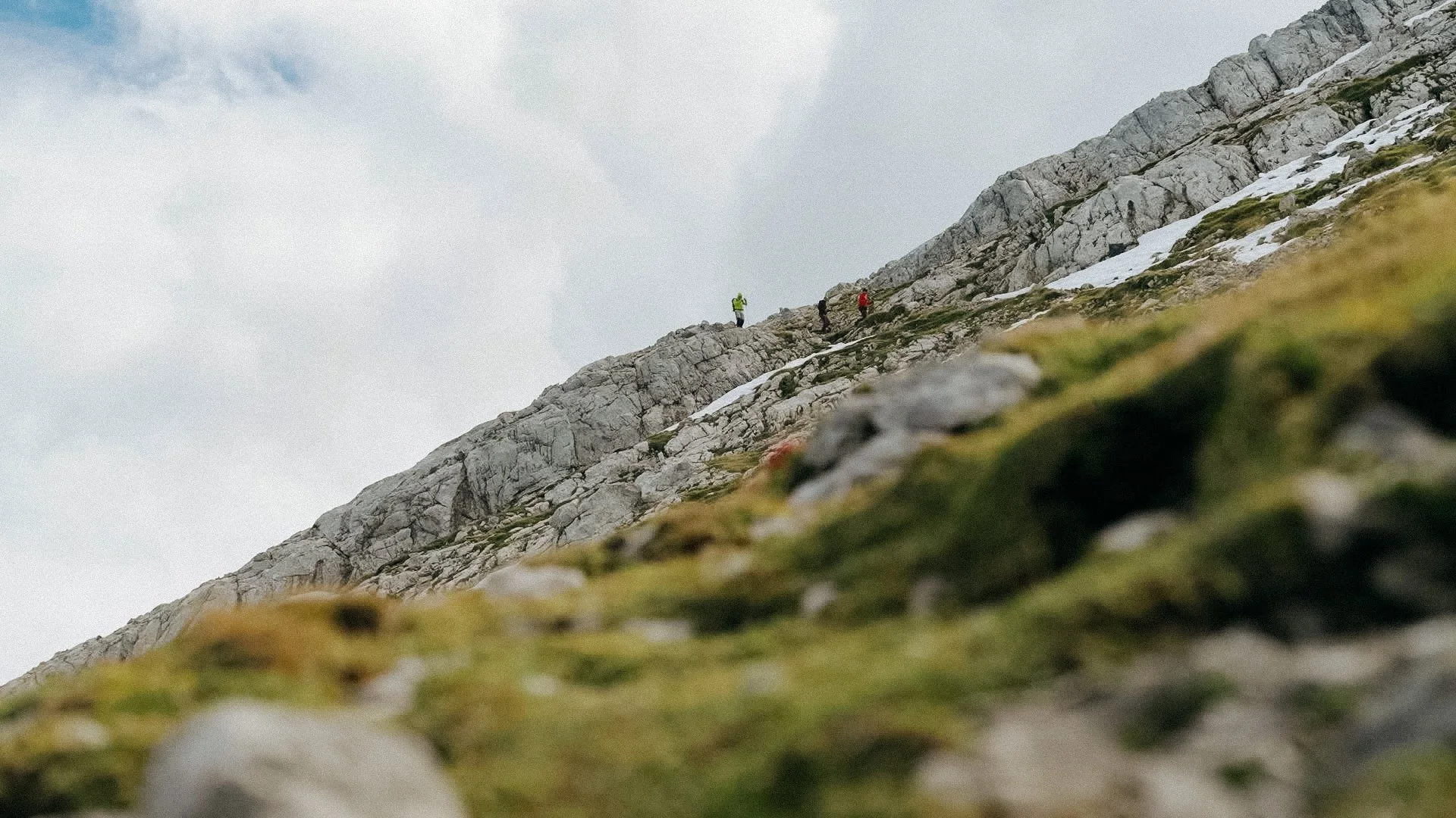 Wanderer auf einem felsigen Bergpfad mit Schneeresten, Blick nach oben auf die Bergkette, Wolken am Himmel.