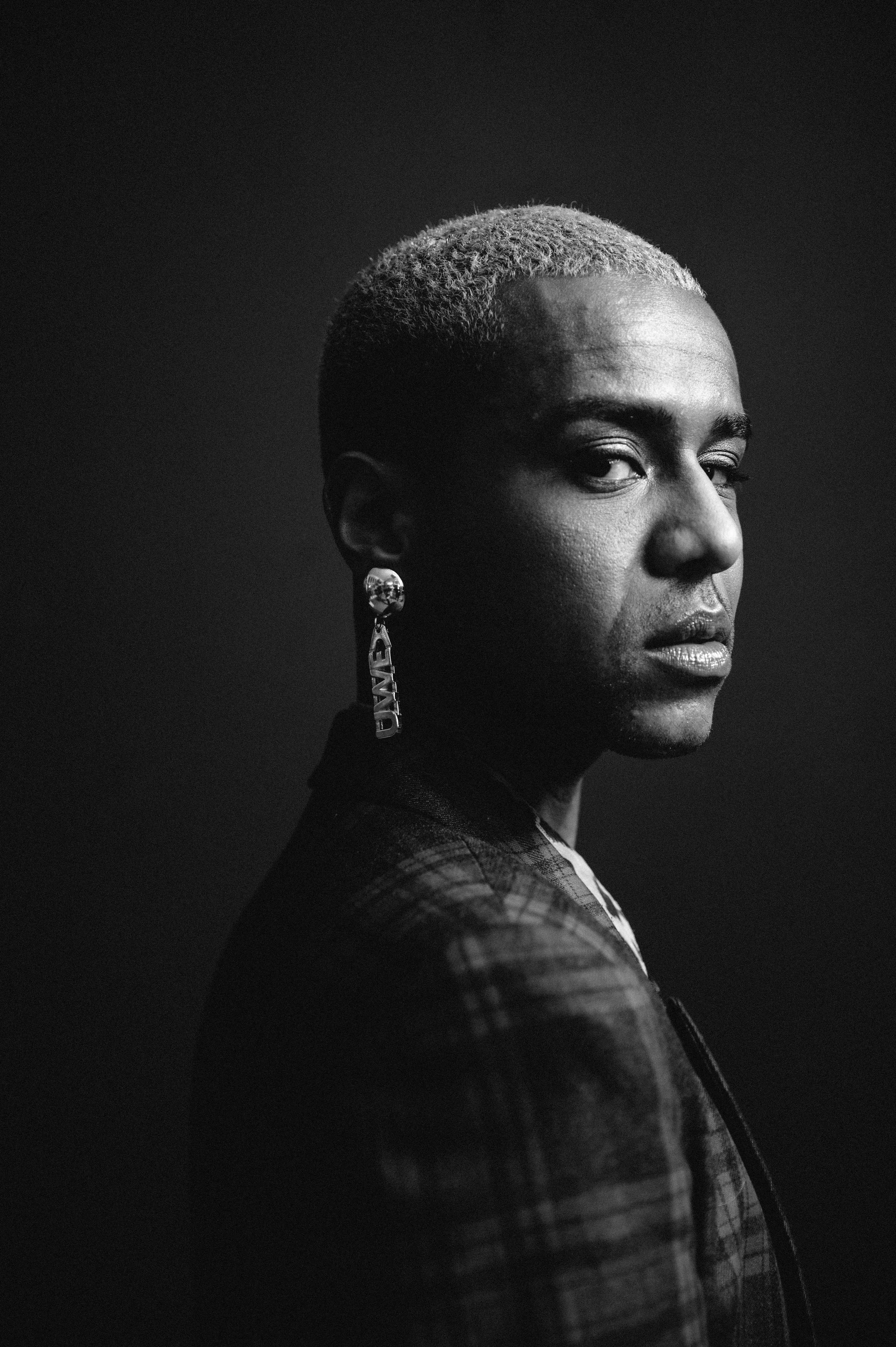 Schwarz-weiß-Porträt einer Person mit kurzes Haar, auffälligem Ohrschmuck und einem ernsthaften Gesichtsausdruck, Blick zur Seite.