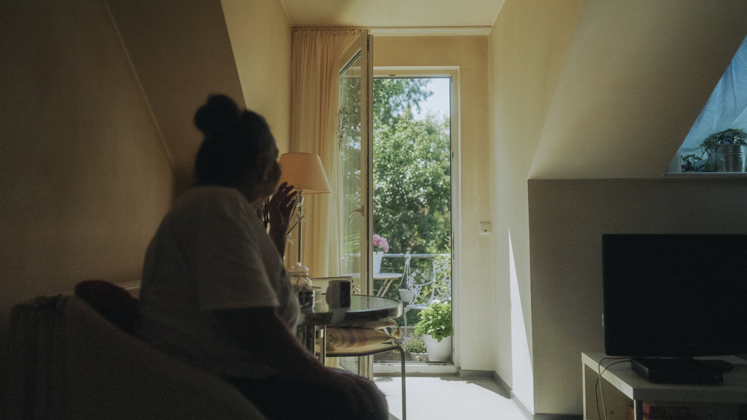 Frau sitzt im dunklen Raum, schaut durchs offene Fenster auf einen Balkon mit Pflanzen und Blick auf grüne Bäume.