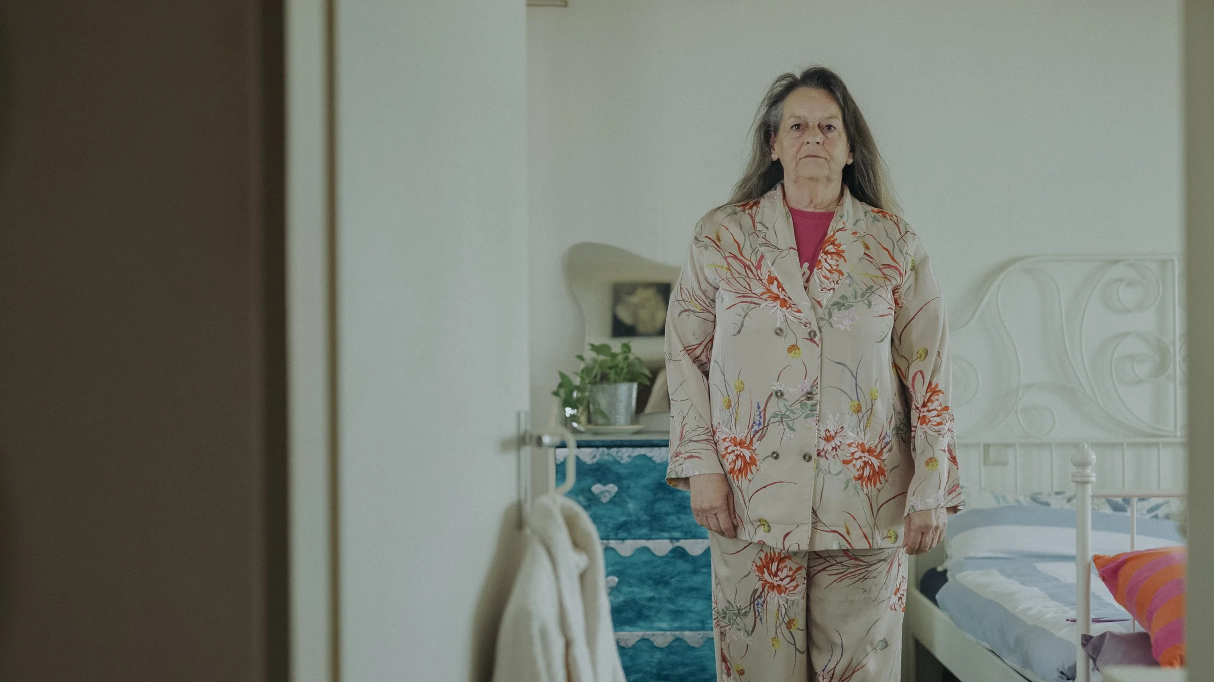 Ältere Frau steht in einem Schlafzimmer, trägt eine helle, bunte Blümchen-Pyjama, wirkt ernst, im Hintergrund Bett und Nachttisch mit Pflanze.