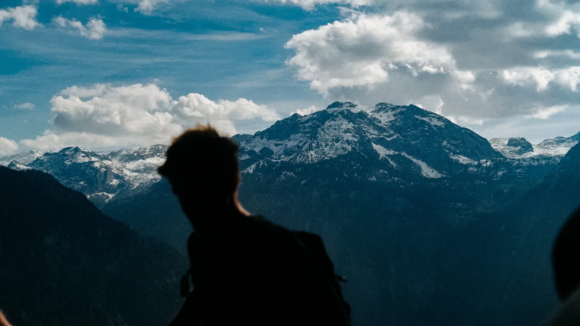 Silhouette einer Person im Vordergrund mit Blick auf eine Berglandschaft mit schneebedeckten Bergen und bewölktem Himmel.