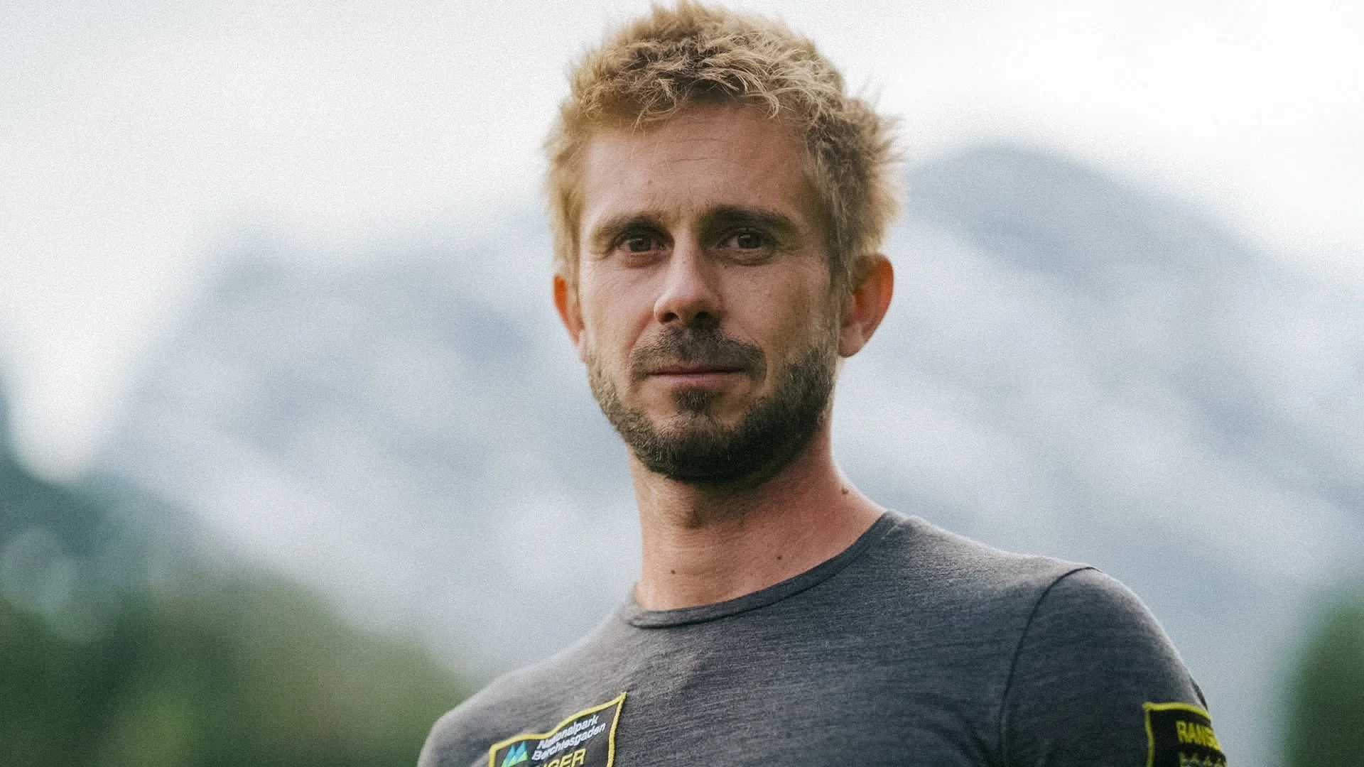 Porträt eines Mannes mit blondem, gelocktem Haar und leichtem Bart vor einer bewölkten, grünen Landschaft.