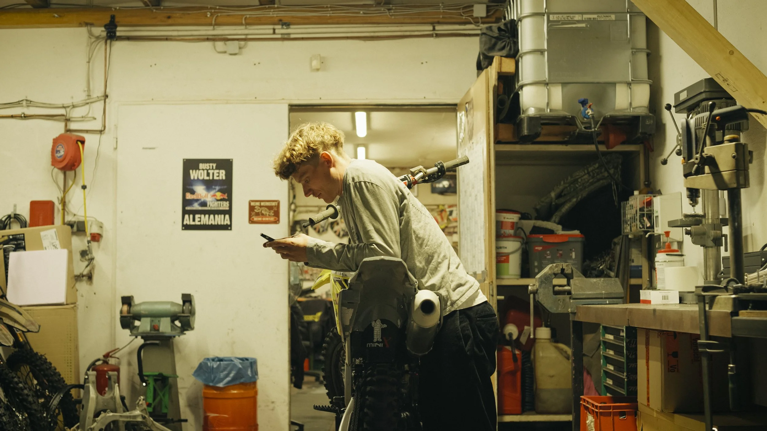 Junge Person in einem Werkstattraum, arbeitet an einem Motorrad und schaut auf sein Smartphone.