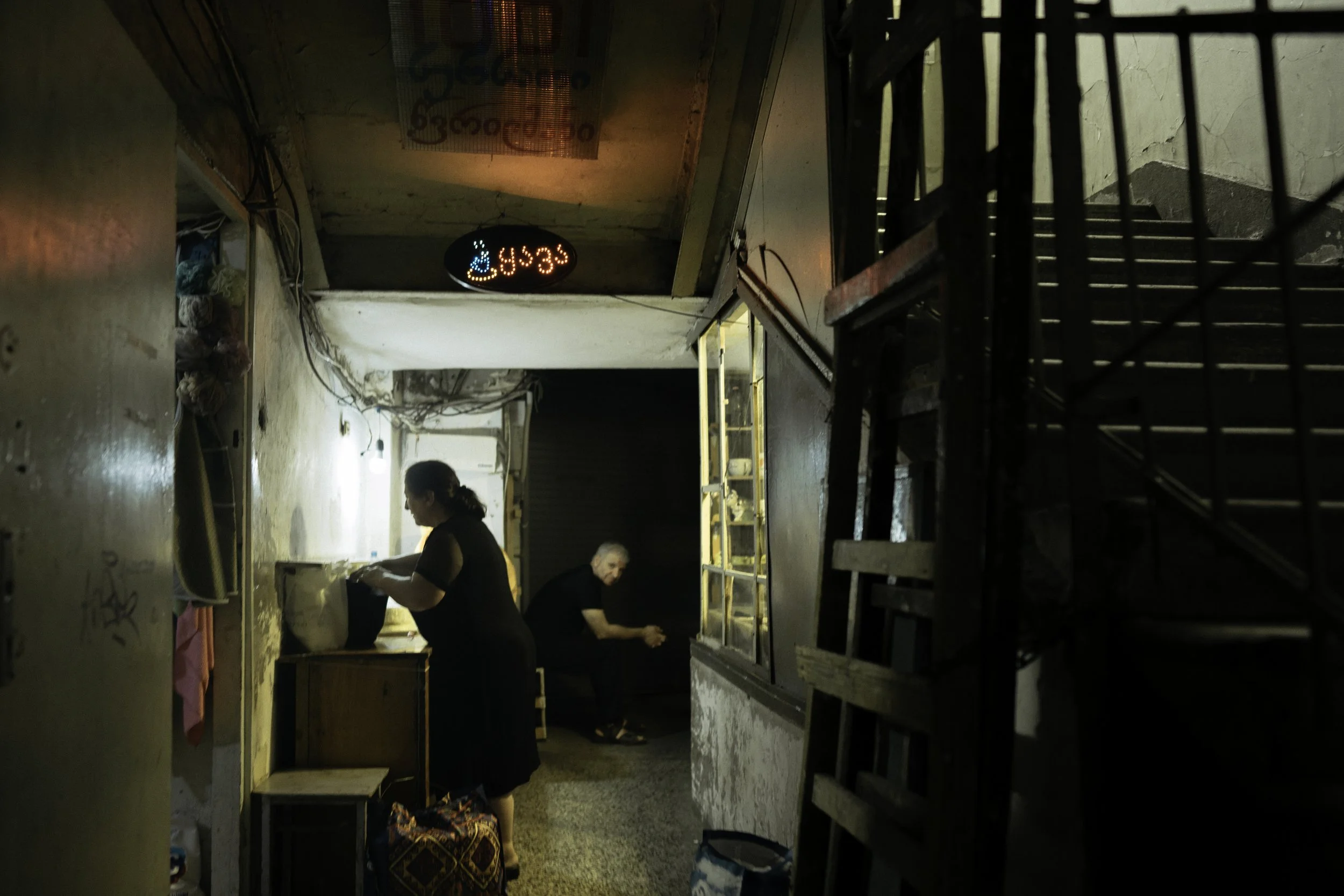 Dunkle Gasse mit zwei Frauen, die an einem kleinen Tisch arbeiten. Über den Frauen eine leuchtende, elektronische Tafel mit georgischer Schrift. Eine Treppe auf der rechten Seite, wenig Licht auf den Wänden.