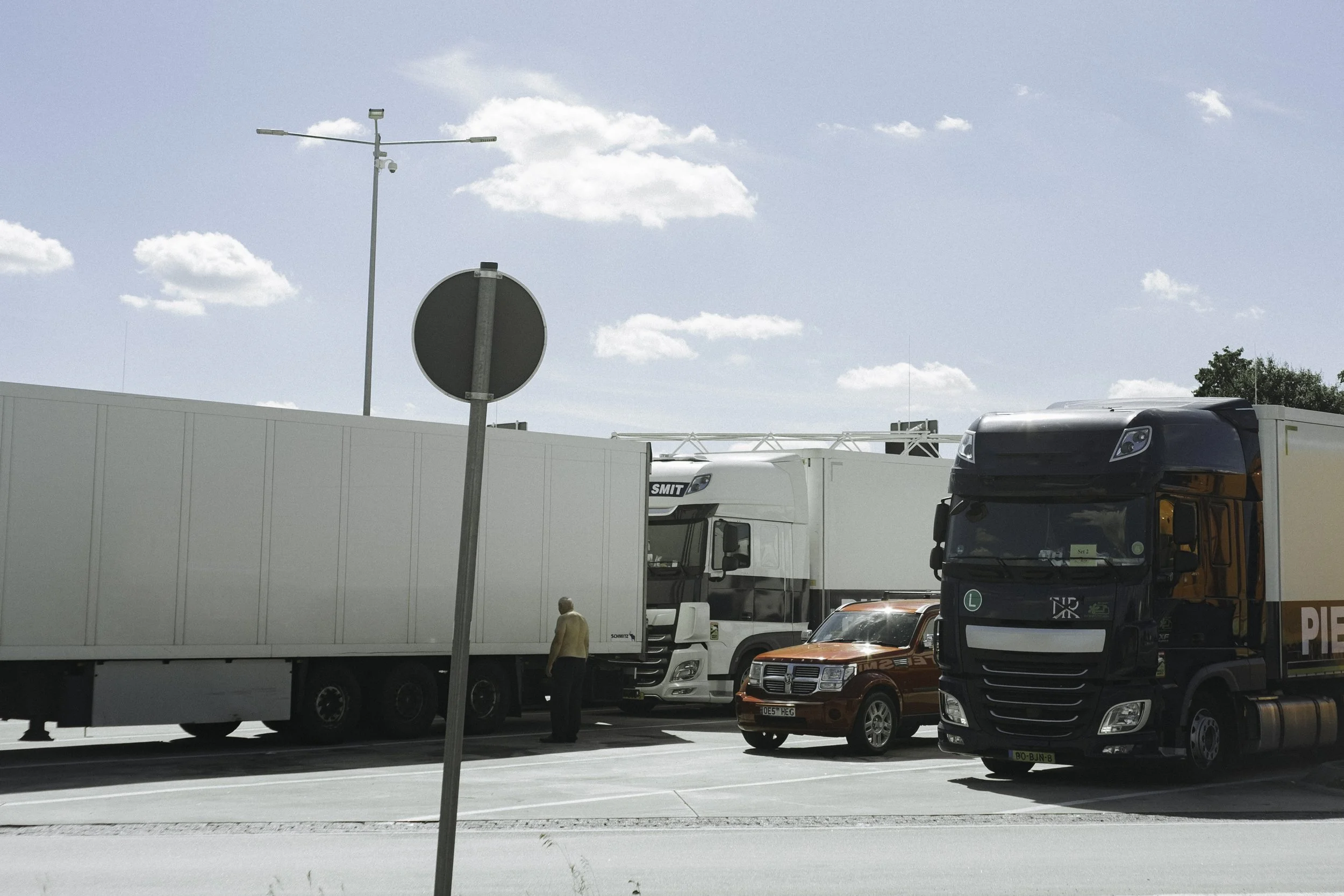 Mehrere große Lastwagen und ein kleiner roter Pkw stehen auf einer Straße. Ein Mann steht neben einem der Lastwagen, während die meisten Fahrzeuge im Schatten stehen. Der Himmel ist blau mit wenigen Wolken.