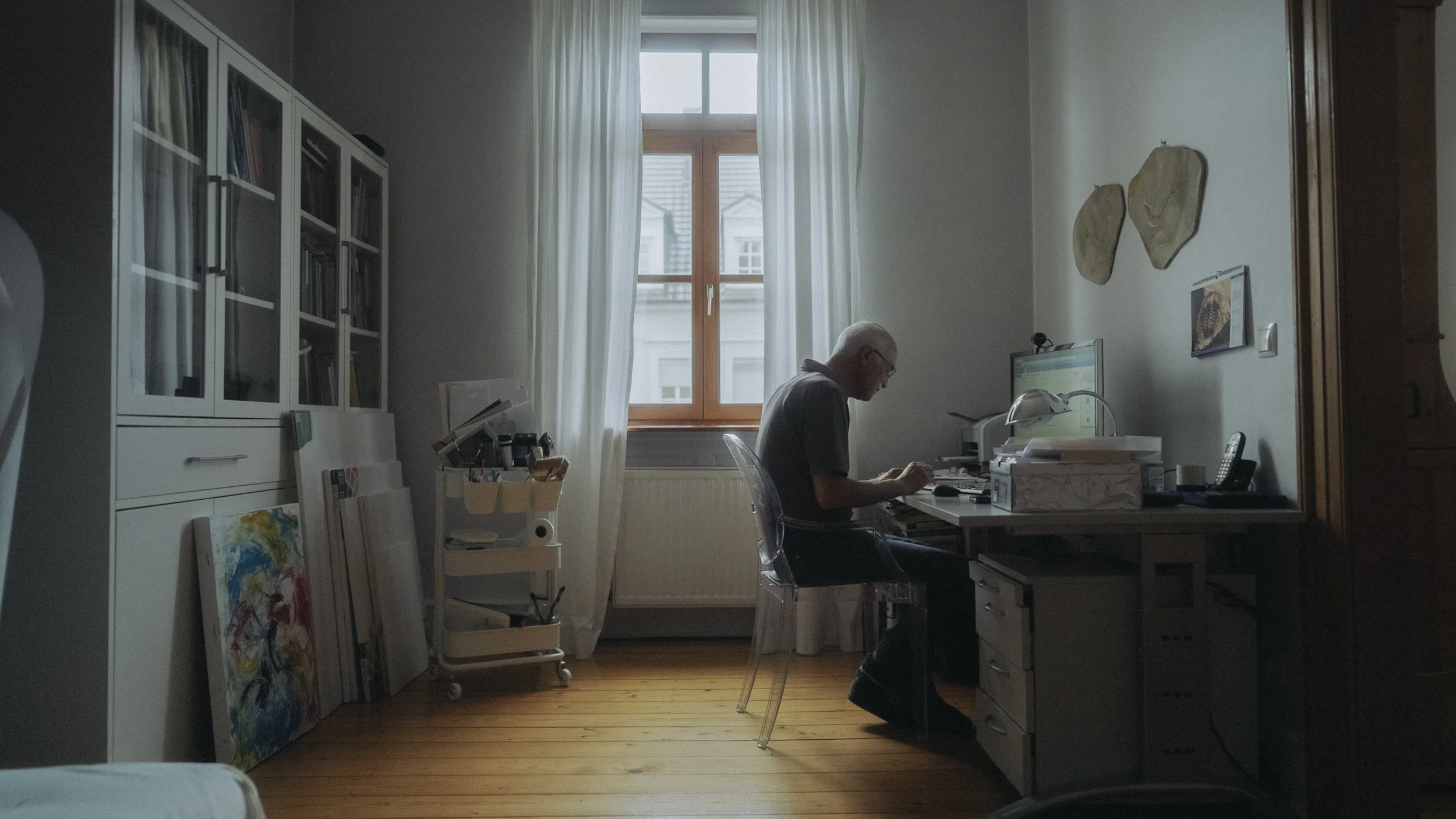 Ein älterer Mann arbeitet an einem Schreibtisch in einem Heimarbeitszimmer mit weißen Wänden, Holzfußboden und einem Fenster mit weißen Vorhängen.