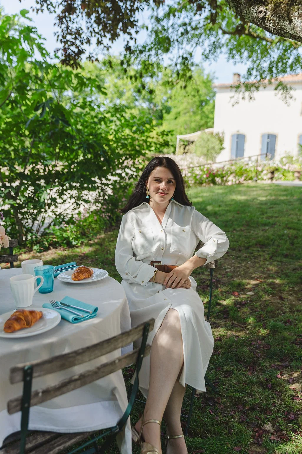 Julie Cousse, une femme habillée en blanc assise à une table de jardin avec des croissants, des tasses et des couverts, dans un cadre verdoyant en plein air.
