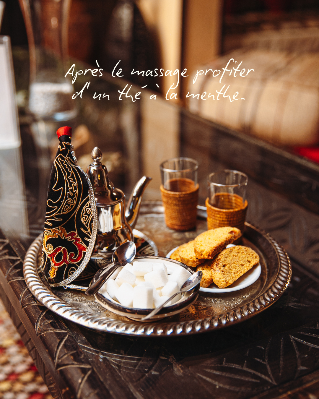 Tasse de thé à la menthe avec des biscuits, du sucre blanc, et une théière sur un plateau en argent, décoré avec un tissu brodé, sur une table en bois.