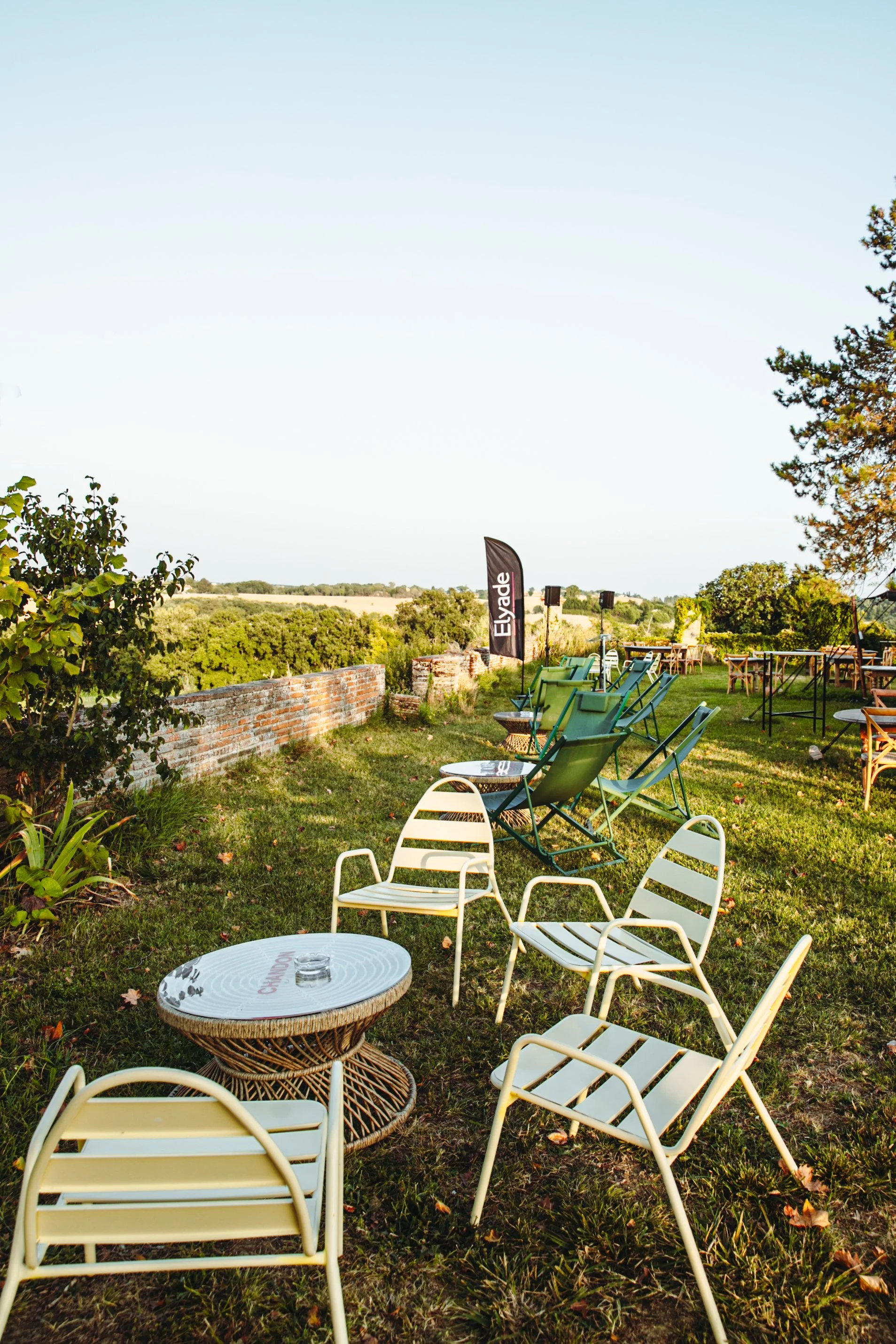 Terrasse extérieur avec plusieurs chaises en métal, tables rondes, espace vert, ciel clair, environnement calme et ensoleillé