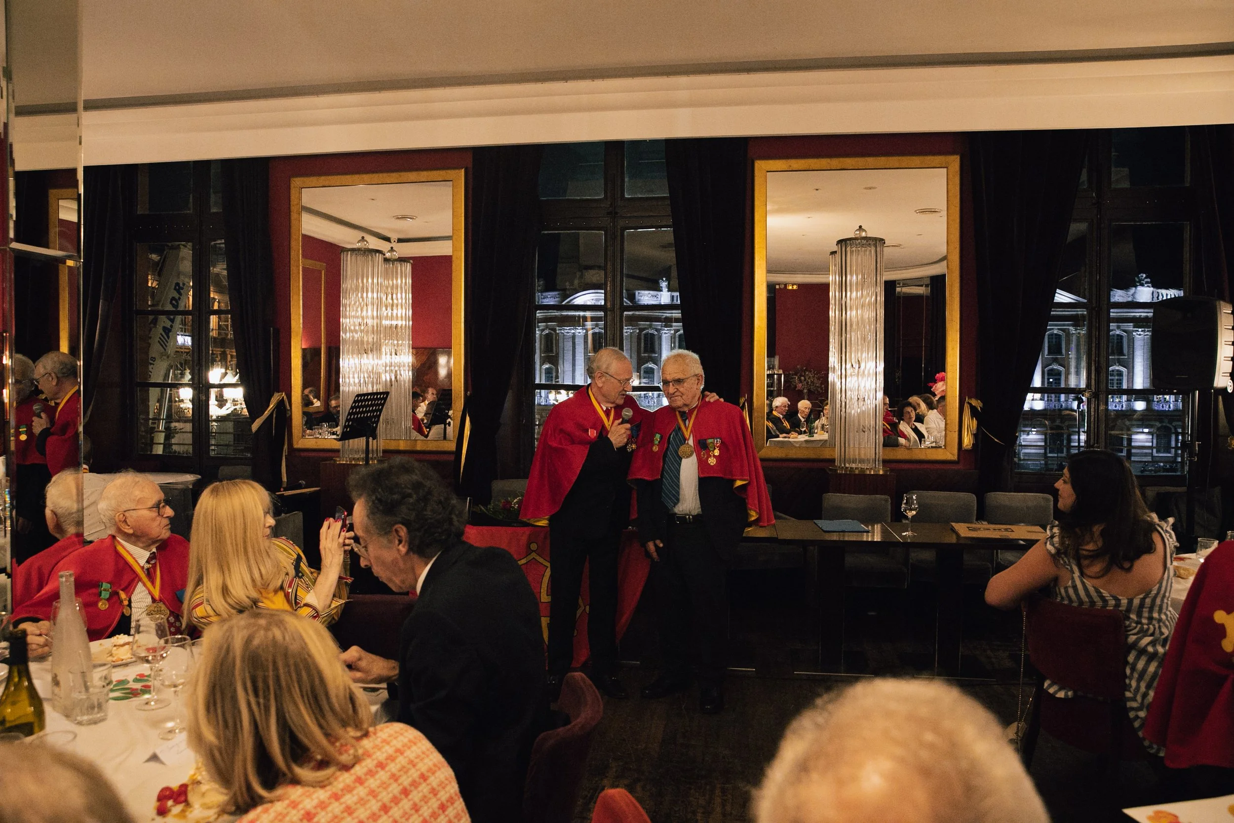 Un banquet ou cérémonie avec des personnes âgées en costume rouge, certains portant des médailles. Deux hommes se parlent, l'un tient un micro. La salle est décorée élégamment avec des miroirs, des lumières et des draperies noires.