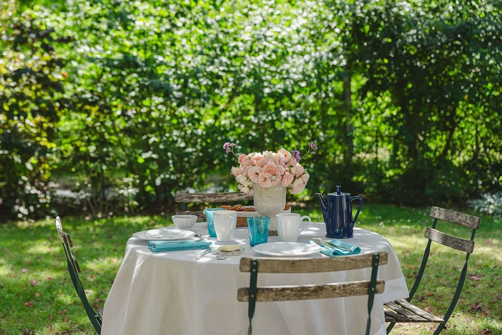 Table de jardin avec une nappe blanche, fleurs roses en vase, vaisselle bleue et blanche, et chaises en bois dans un jardin ensoleillé