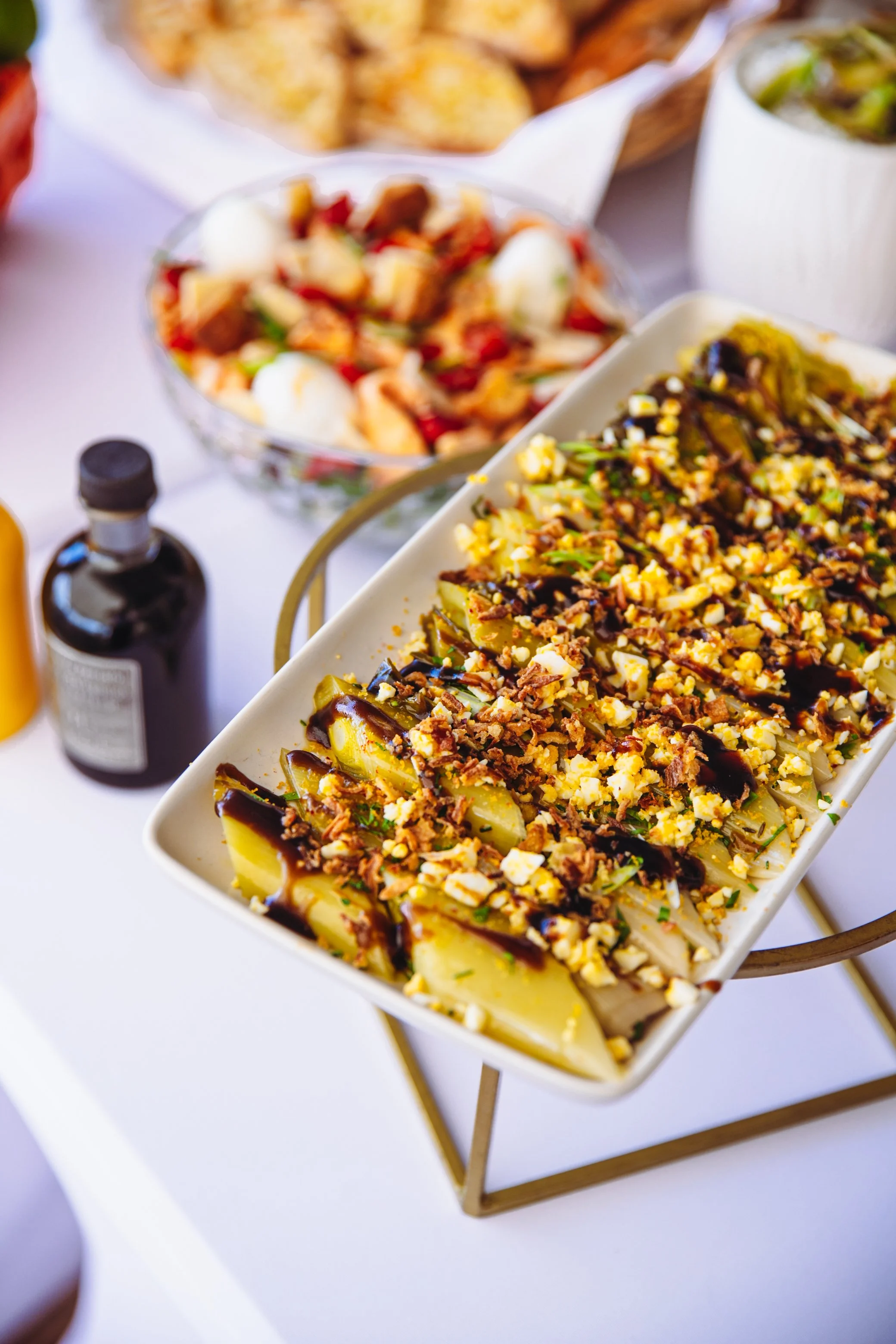 Plat de lamelles de céleri rave garnies de noix hachées, de sauce balsamique et d'herbes, présenté dans un plat blanc sur un support en métal, avec des salades et des condiments en arrière-plan.