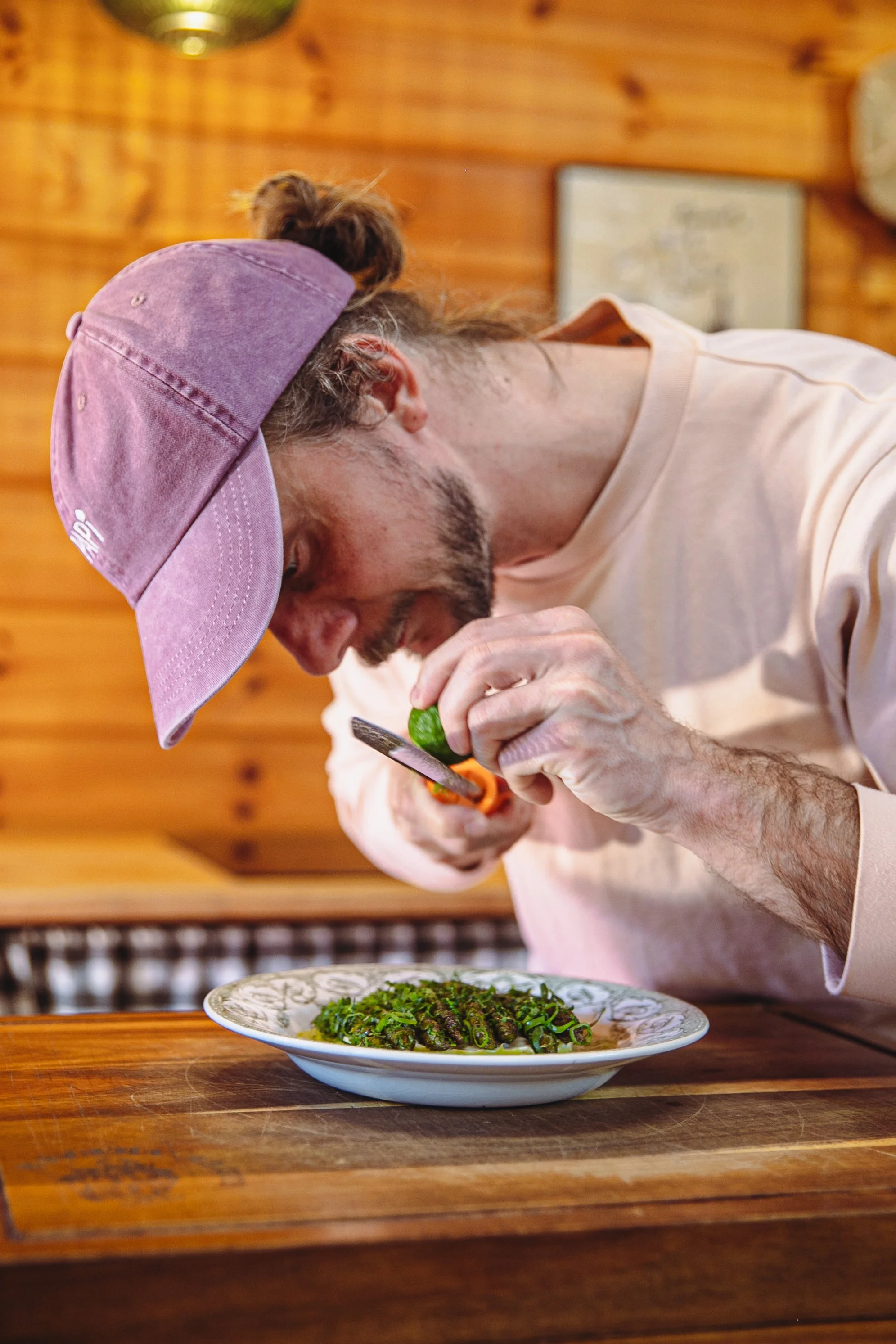 Un homme avec une casquette violette et une barbe, en train de garnir un plat d'asperges avec des herbes, dans une pièce avec murs en bois.
