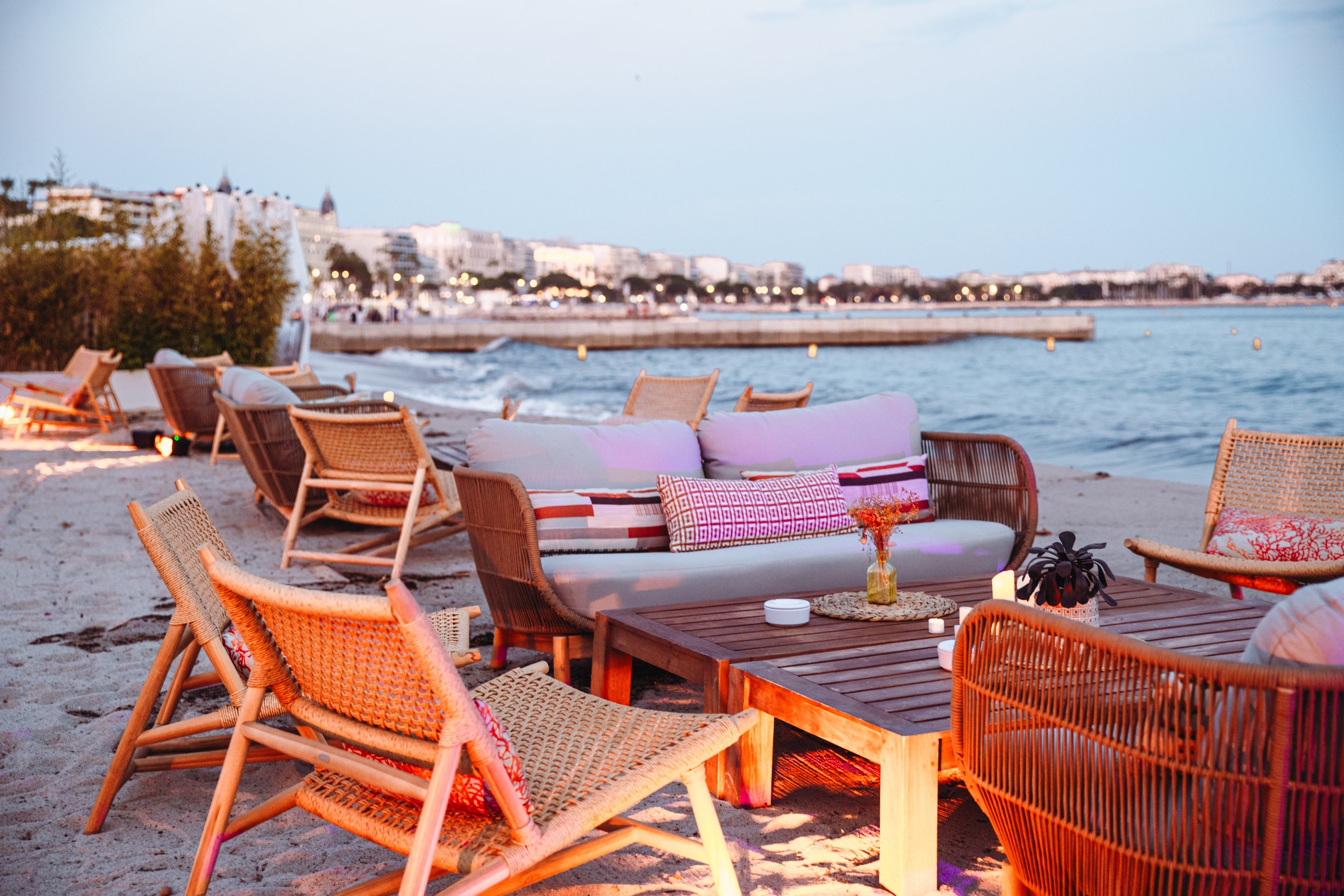 Coin lounge sur la plage avec fauteuils en bois, canapé avec coussins, table en bois, bougies, décor sobre, vue sur l'eau et bâtiments en arrière-plan, ambiance détendue au crépuscule.