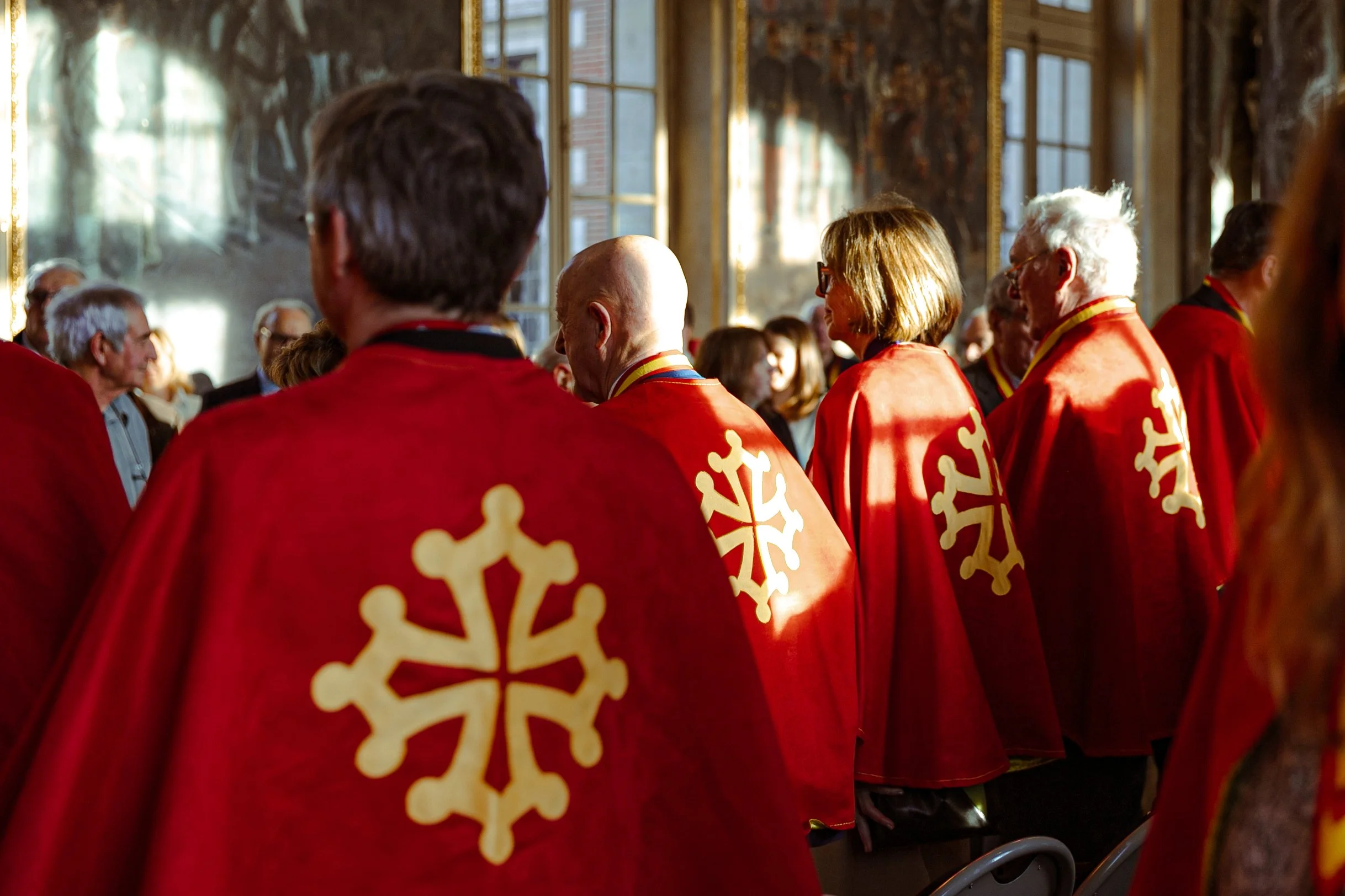 Groupe de personnes portant des capes rouges avec un symbole blanc lors d'une cérémonie à l'intérieur, ambiance lumineuse avec lumière naturelle.