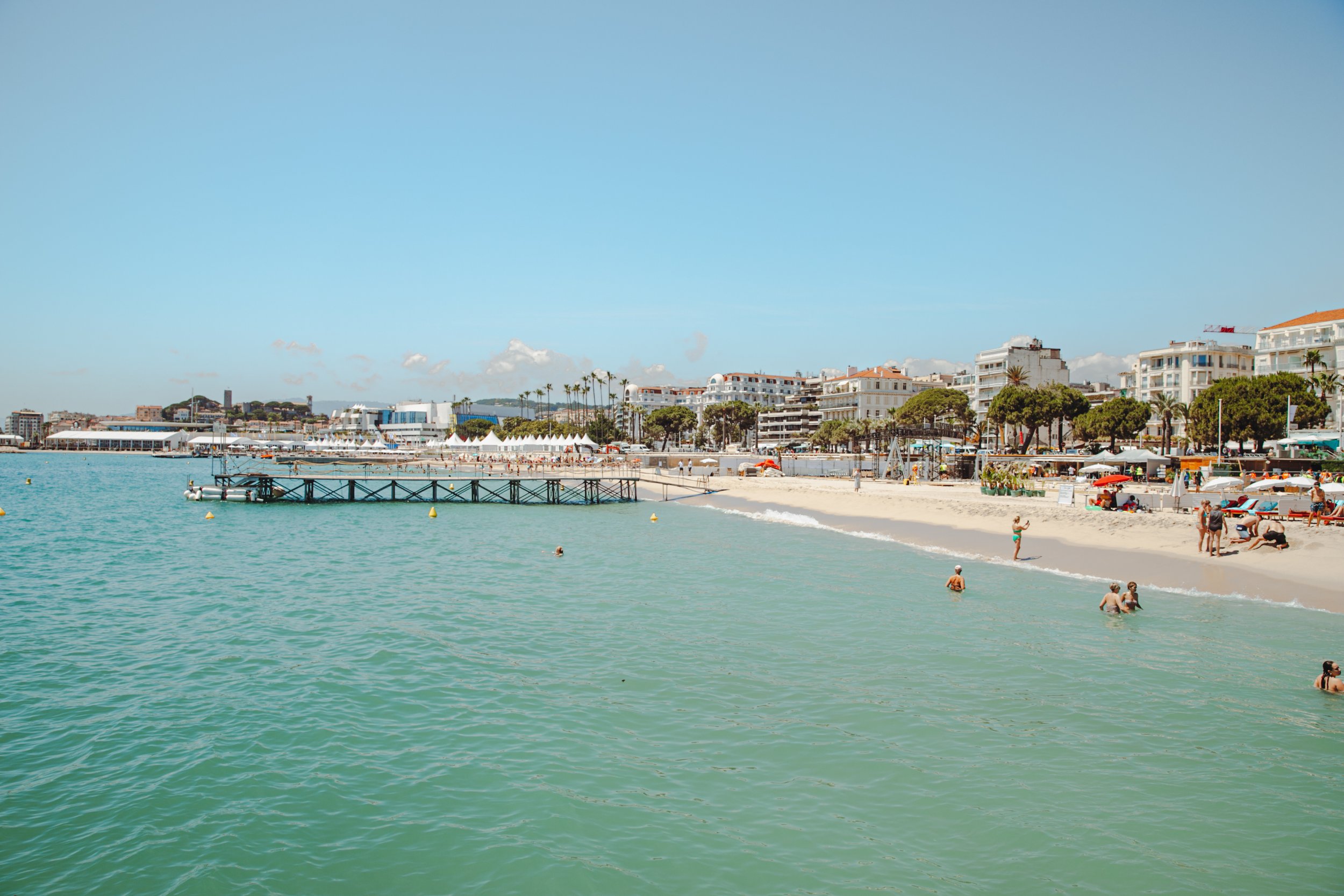 Plage animée avec personnes nageant, se relaxant et profitant du soleil, avec bâtiments modernes et palmiers en arrière-plan sous un ciel clair.