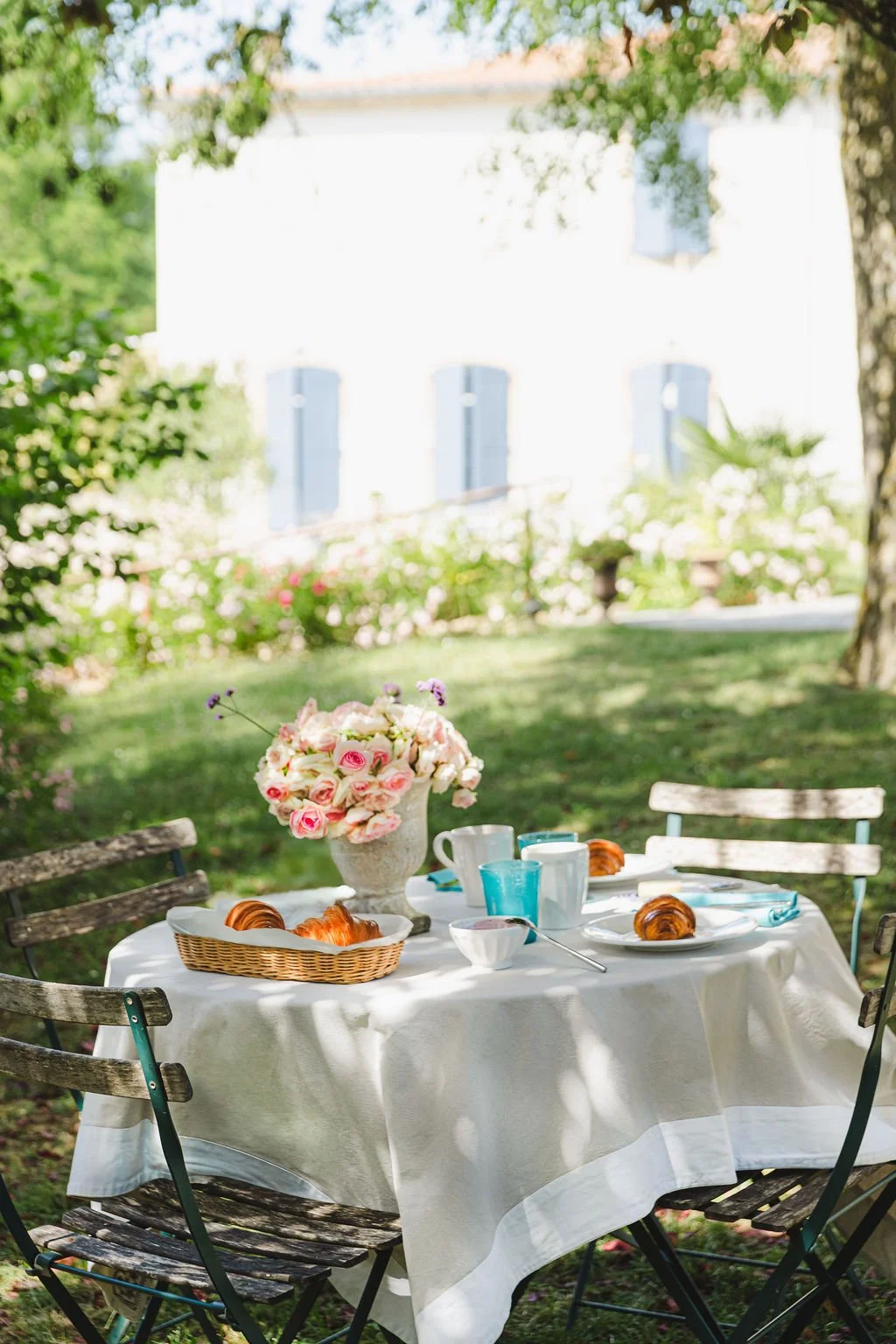 Table de petit déjeuner en plein air avec un vase de fleurs roses, croissants sur des assiettes, tasses, verres bleus et blancs, dans un jardin vert avec une maison en arrière-plan.