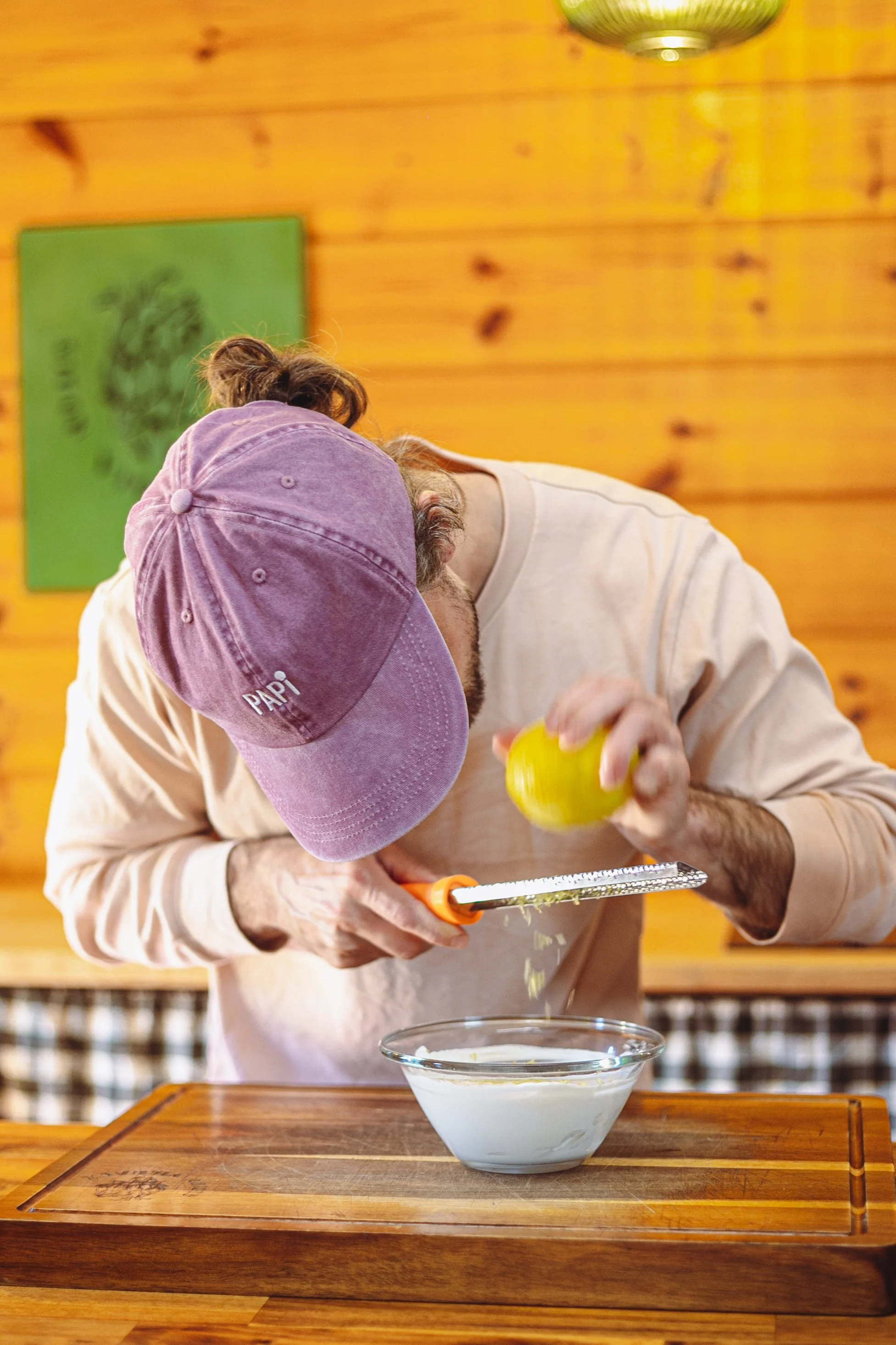 Une personne, portant une casquette violette avec l'inscription 'PAPI', est en train de râper un morceau de citron au-dessus d'un bol blanc contenant ce qui semble être du yaourt ou de la crème. La scène se déroule dans une cuisine chaleureuse avec u