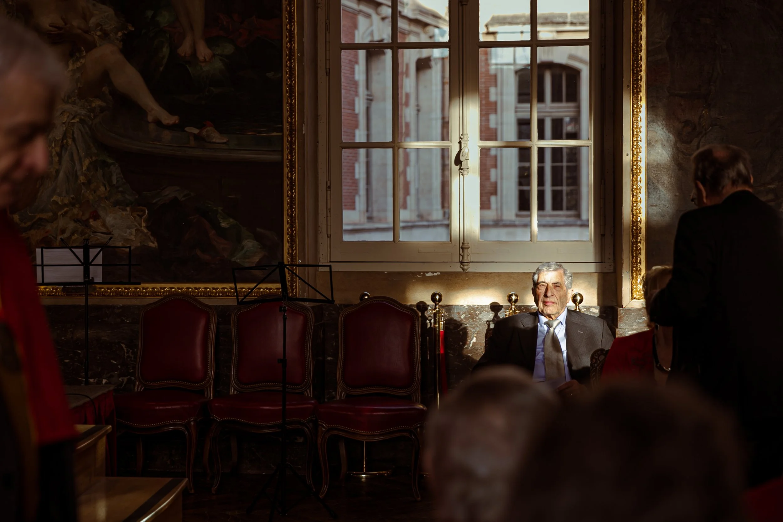 Un homme âgé assis sur un fauteuil doré dans une pièce éclairée par la lumière naturelle d'une fenêtre, entouré de personnes discutant lors d'une réunion ou d'un événement formel