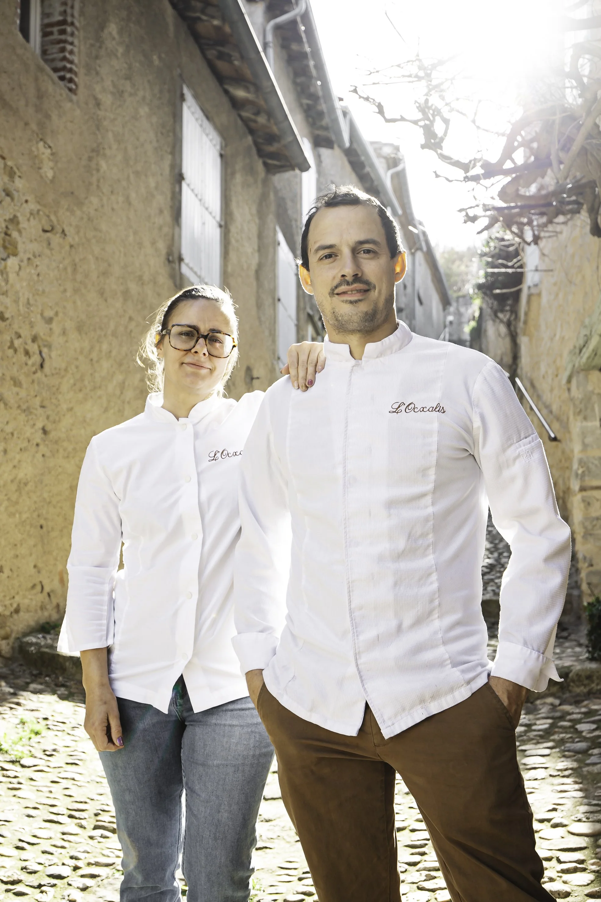 Deux chefs portent des uniformes blancs avec le nom "L'Oceade". Ils se tiennent dans une ruelle pavée en pierre, éclairée par le soleil, avec des maisons anciennes en pierre à l'arrière-plan.
