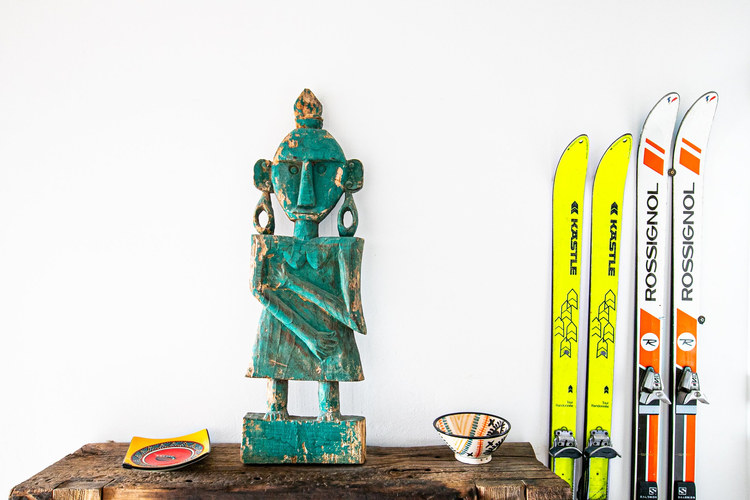 Objets décoratifs et accessoires de sport sur une table en bois contre un mur blanc, notamment une sculpture en bois peinte en turquoise, deux petits plateaux avec motifs, un bol, et des skis assis verticalement.