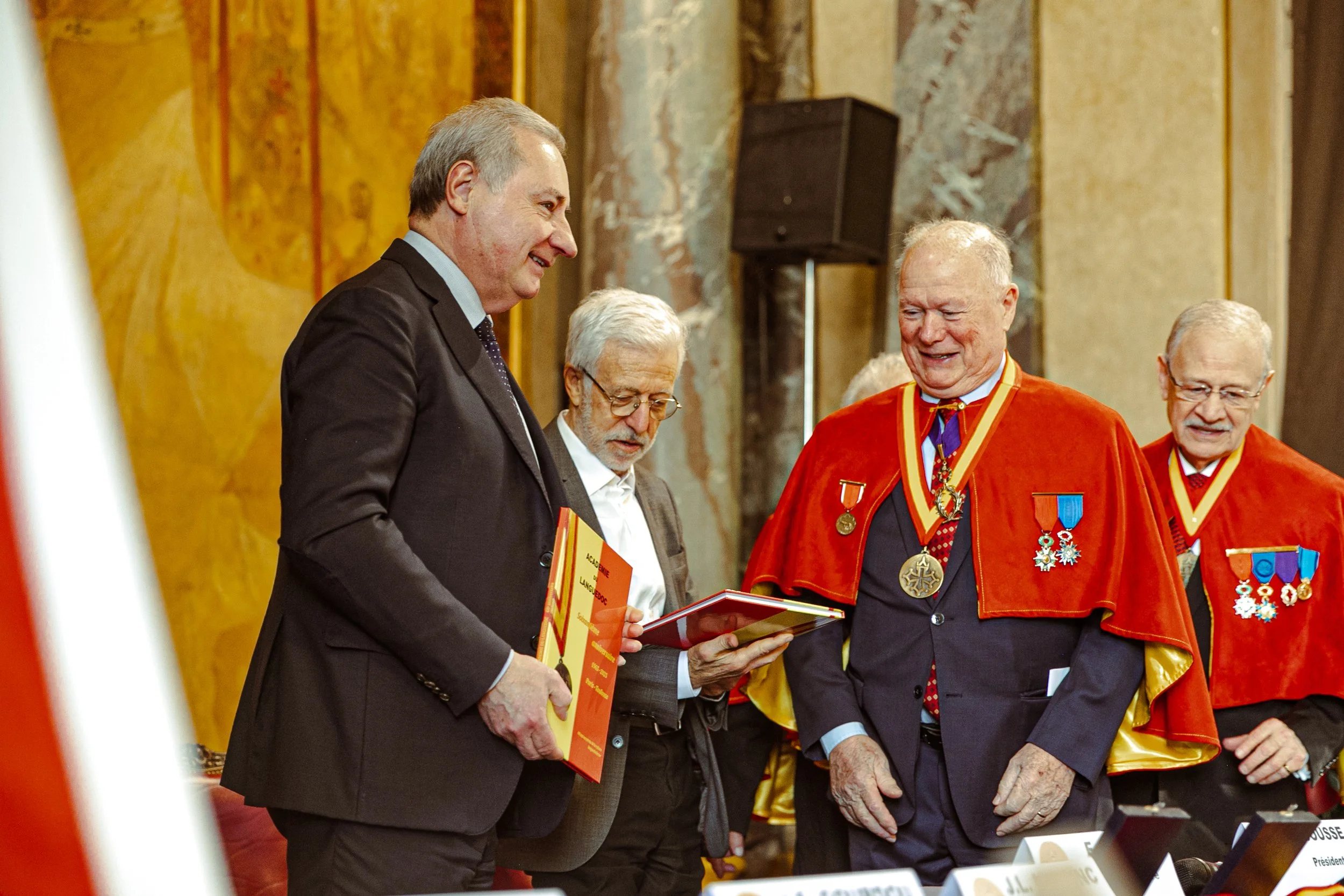 Cinq hommes lors d'une cérémonie officielle, deux portent des médailles et des manteaux rouges, un montre une médaille, ils sourient.