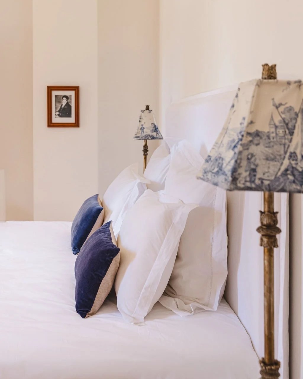 Un lit fait avec plusieurs oreillers blancs et deux oreillers gris et violet, avec deux lampes de chevet à lampes en tissu à motifs bleus, et un cadre photo en bois sur le mur blanc derrière.