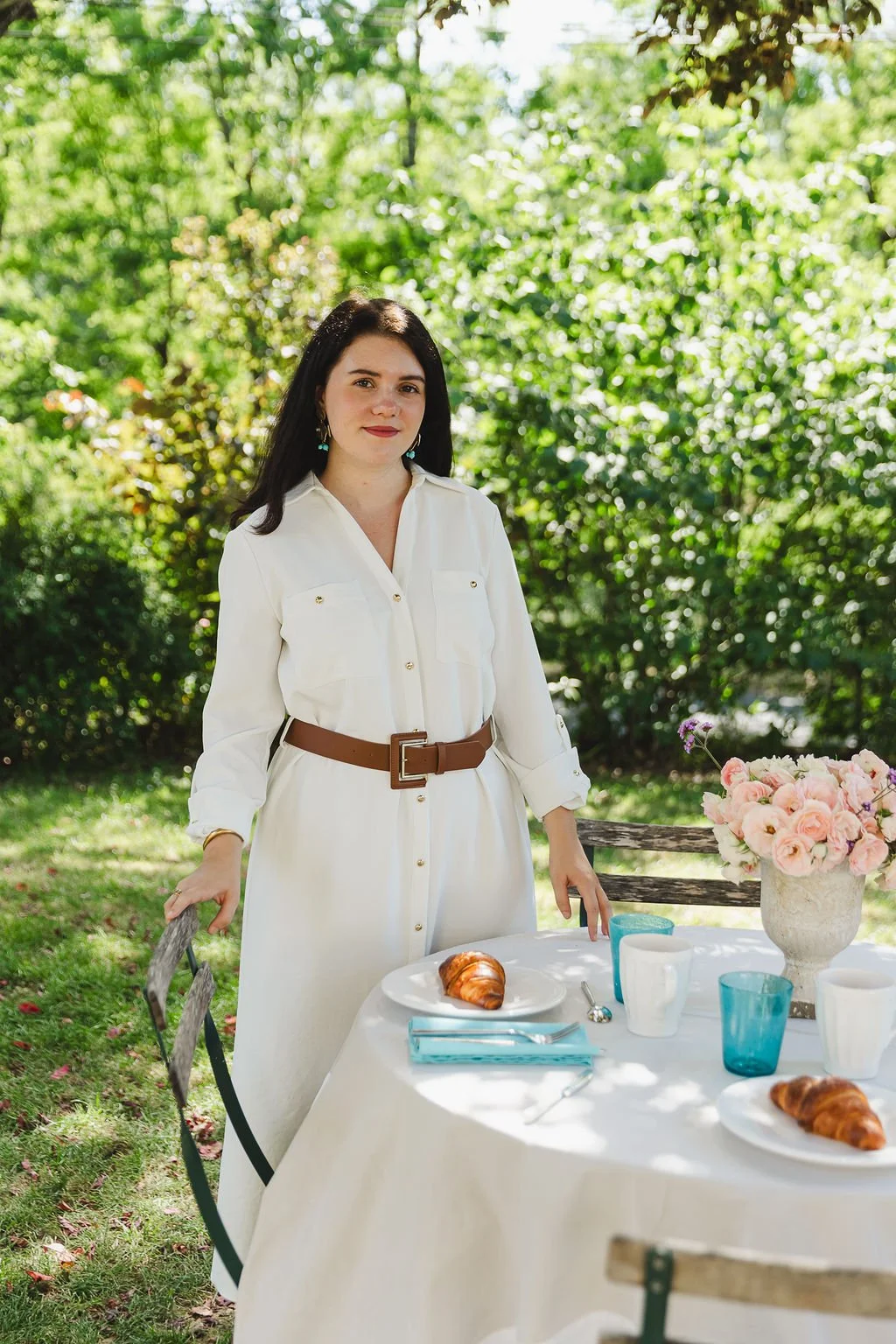 Julie Cousse, une femme debout à une table de pique-nique en plein air, avec un bouquet de fleurs roses et blancs, des croissants, des tasses et des verres colorés, dans un jardin verdoyant.