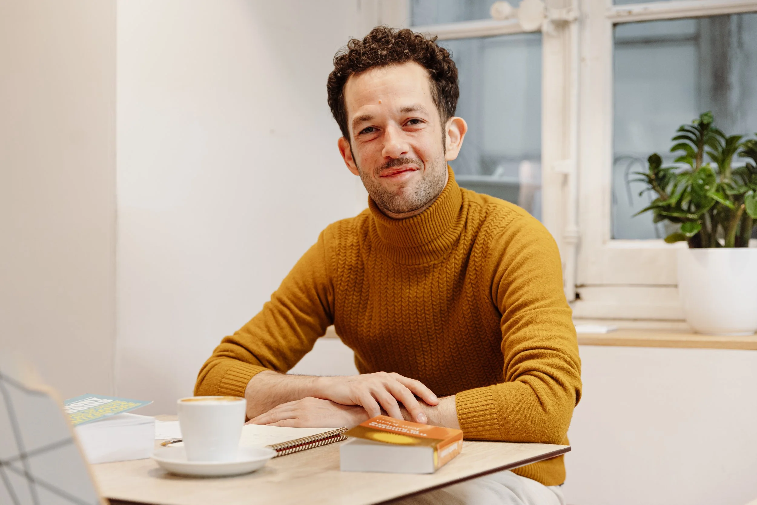 Un homme souriant, portant un pull de couleur marron, assis à une table avec une tasse de café, un livre, et des papiers, près d'une fenêtre avec une plante en pot.