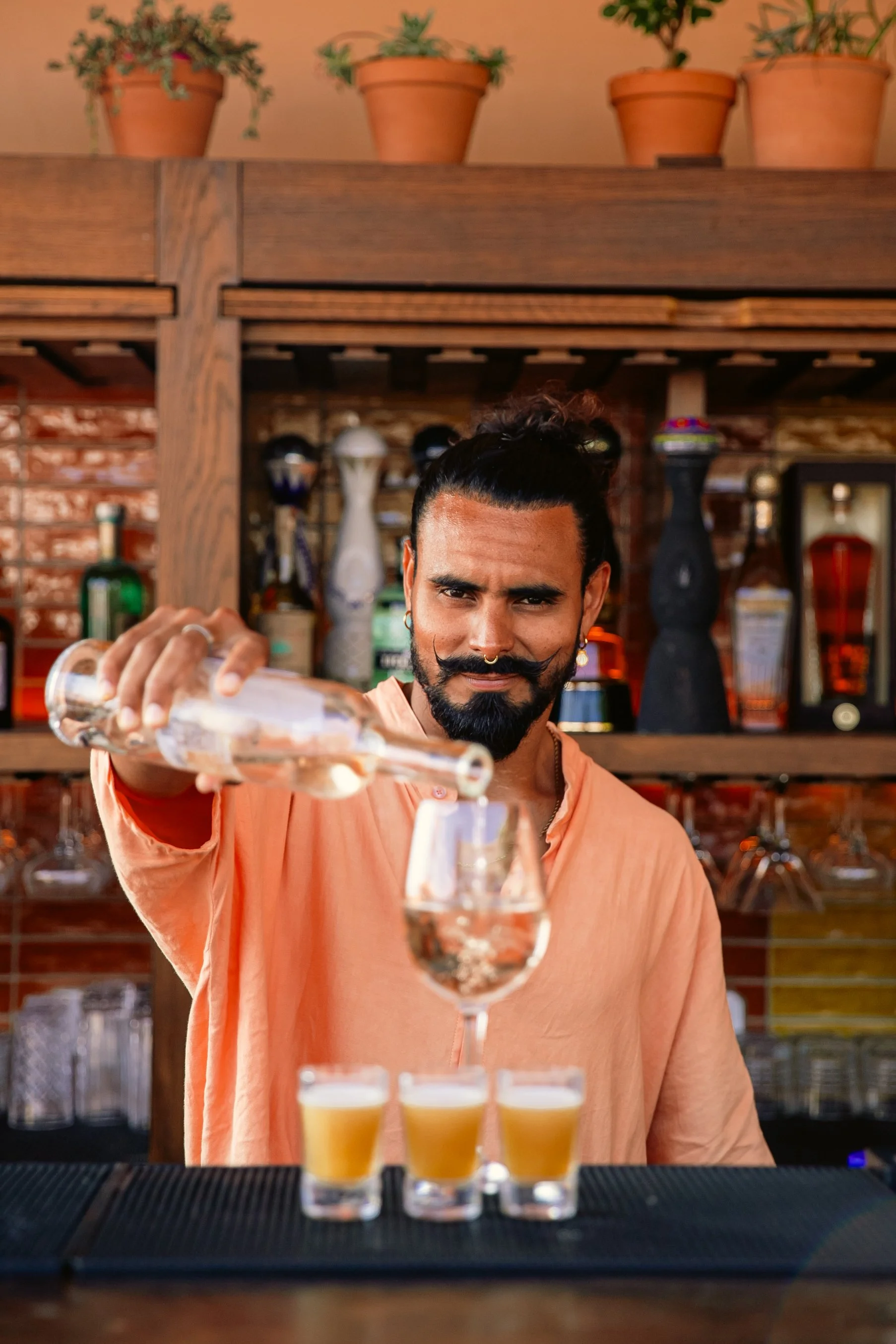 Barman avec un tatouage au bras, portant une robe orange, verse une boisson dans un verre à cocktail, avec quatre petits verres de bière devant lui, dans un bar avec étagères en bois et bouteilles en arrière-plan.