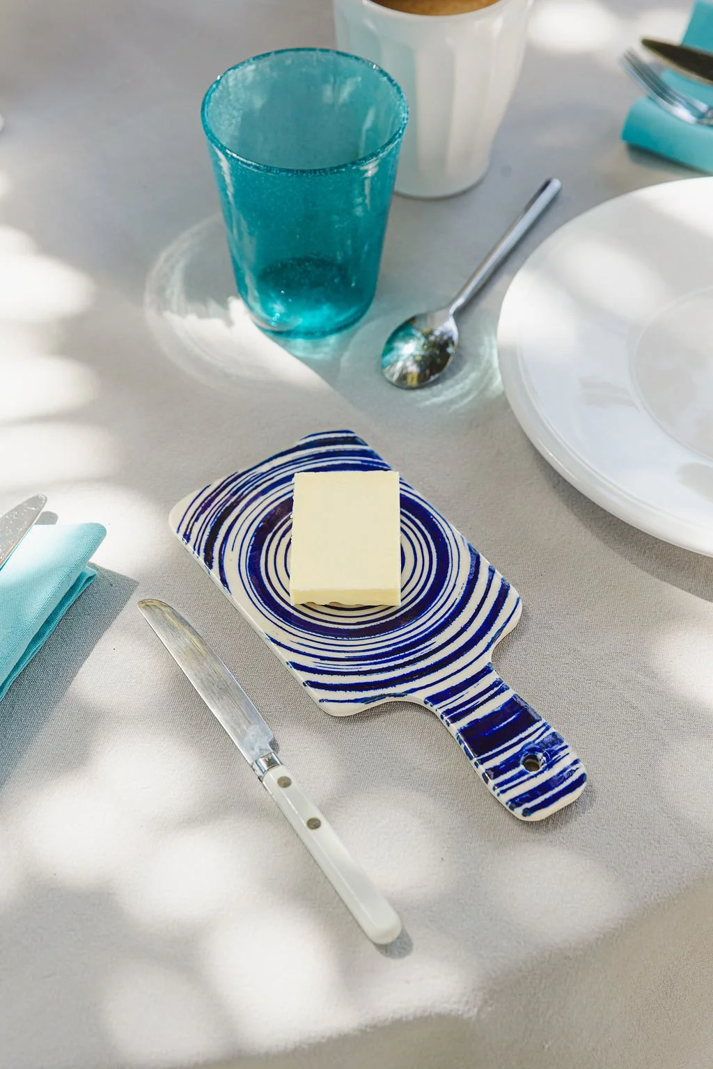 Une table avec une assiette blanche, un verre de boisson bleue, une cuillère, un couteau, un beurre sur une planche à découper en céramique bleue et blanche, un bol blanc et une serviette bleue.