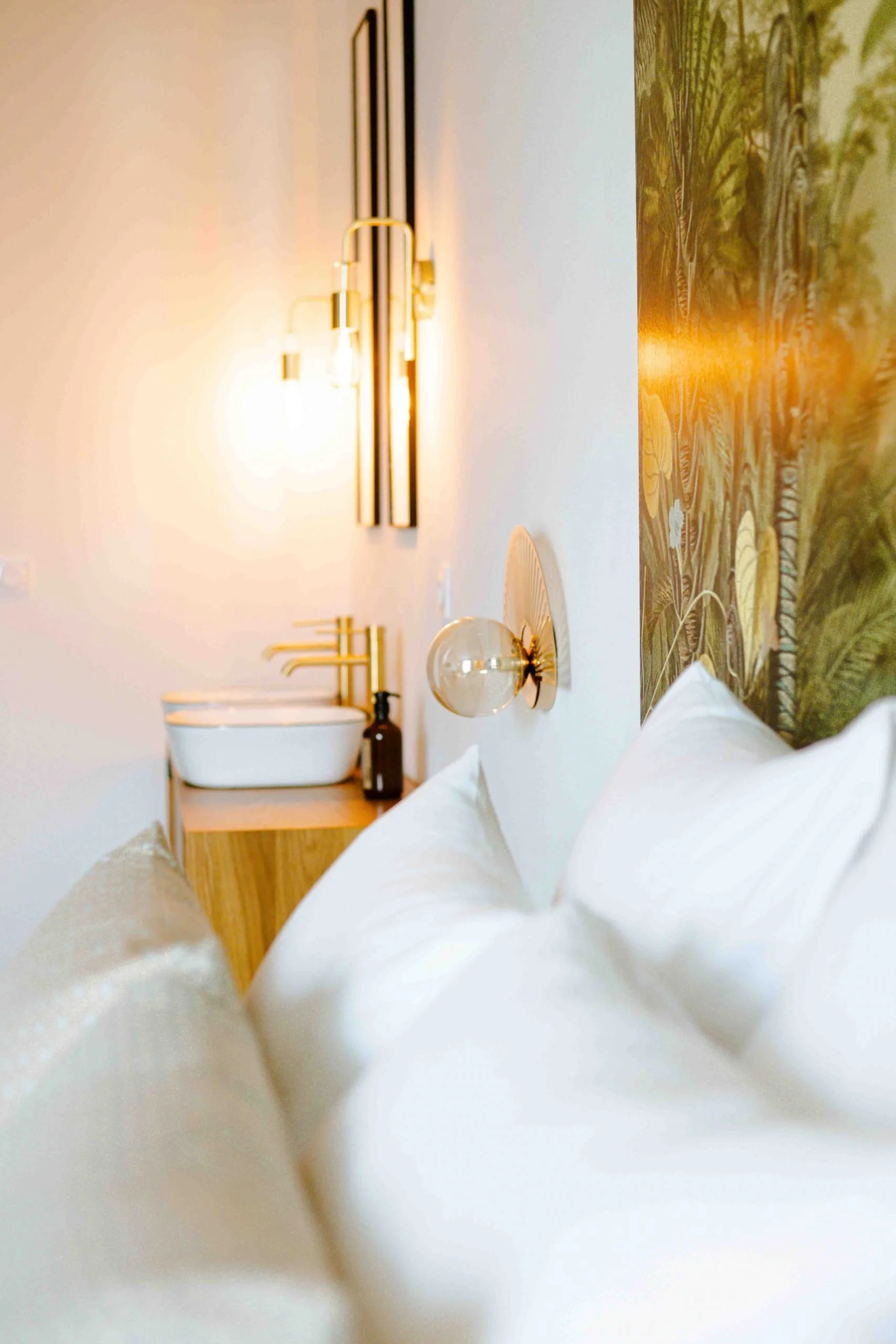 Vue d'un lit avec des oreillers blancs, un mur avec un éclairage moderne et une tête de lit avec motif végétal, à côté d'une petite table de nuit avec un lavabo blanc et une bouteille.