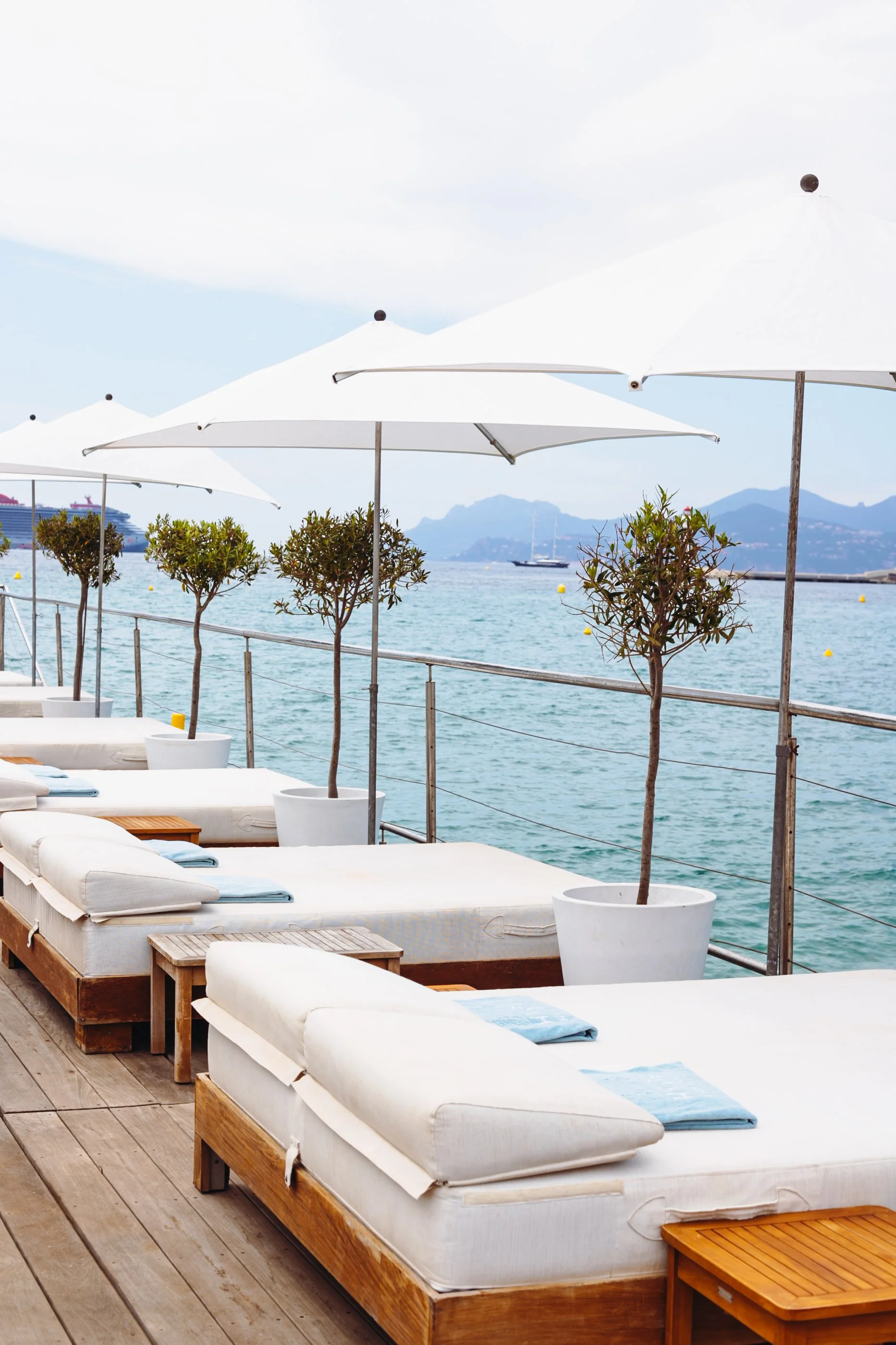 Zones détente avec matelas en bois, parasols blancs, petits arbres en pots le long d'une mer calme, montages en arrière-plan.