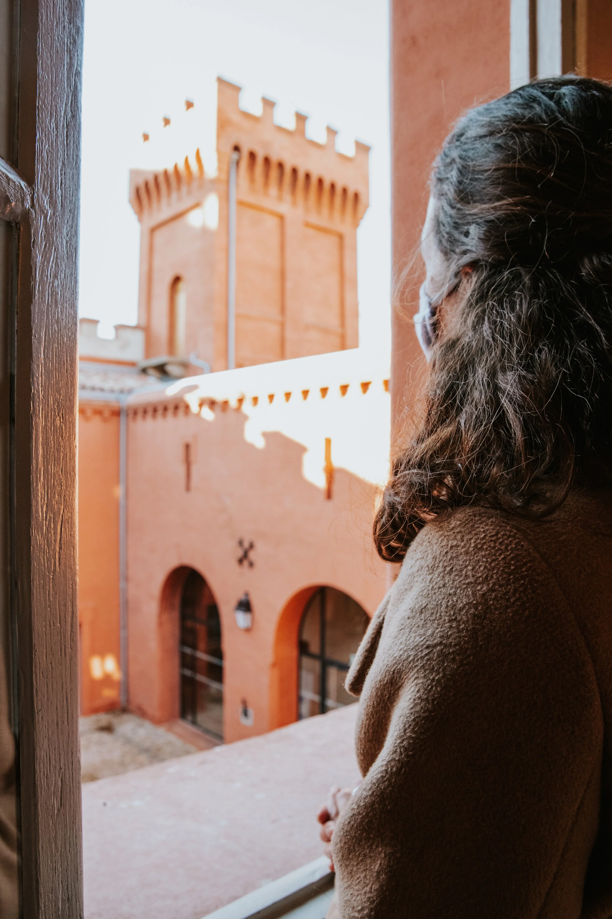 Une femme regarde par une fenêtre vers un bâtiment en briques rouges avec une tour. Elle porte un manteau marron et un masque.