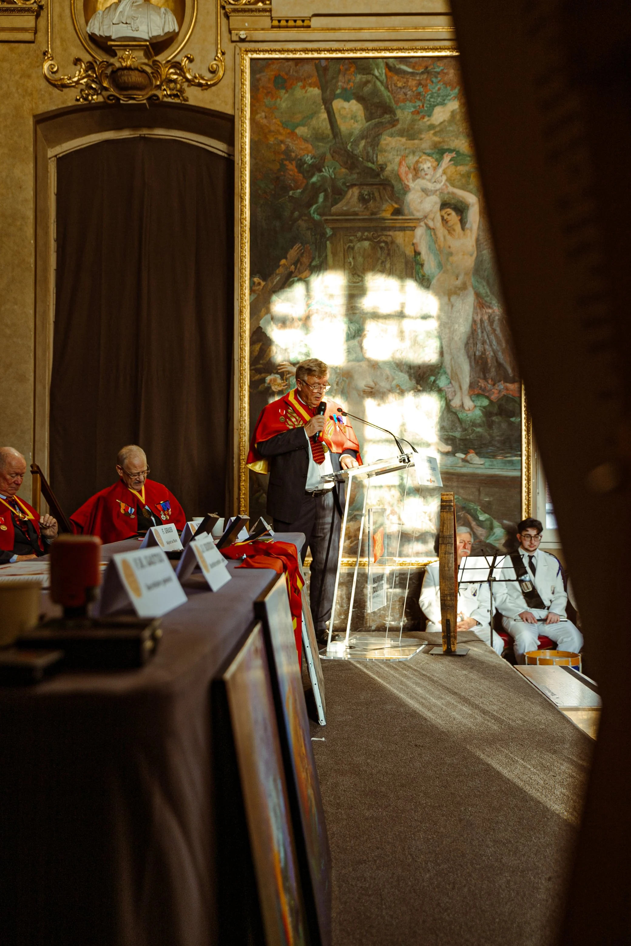 Un homme parle lors d'une cérémonie ou d'une conférence, portant une veste avec des décorations rouges, devant un grand tableau de style classique, avec plusieurs personnes assises portant des robes ou des uniformes, dans une salle richement décorée.