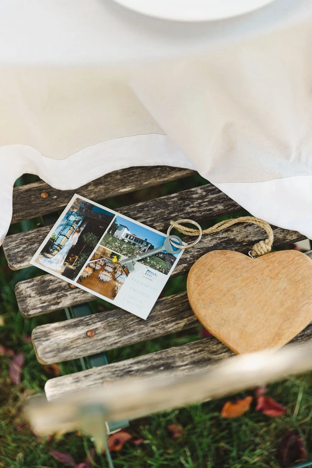 Une photo prise à l'extérieur montrant une petite table en bois avec une carte postale, une clé avec une bague et un cœur en bois. La scène est partiellement couverte d'un tissu blanc.