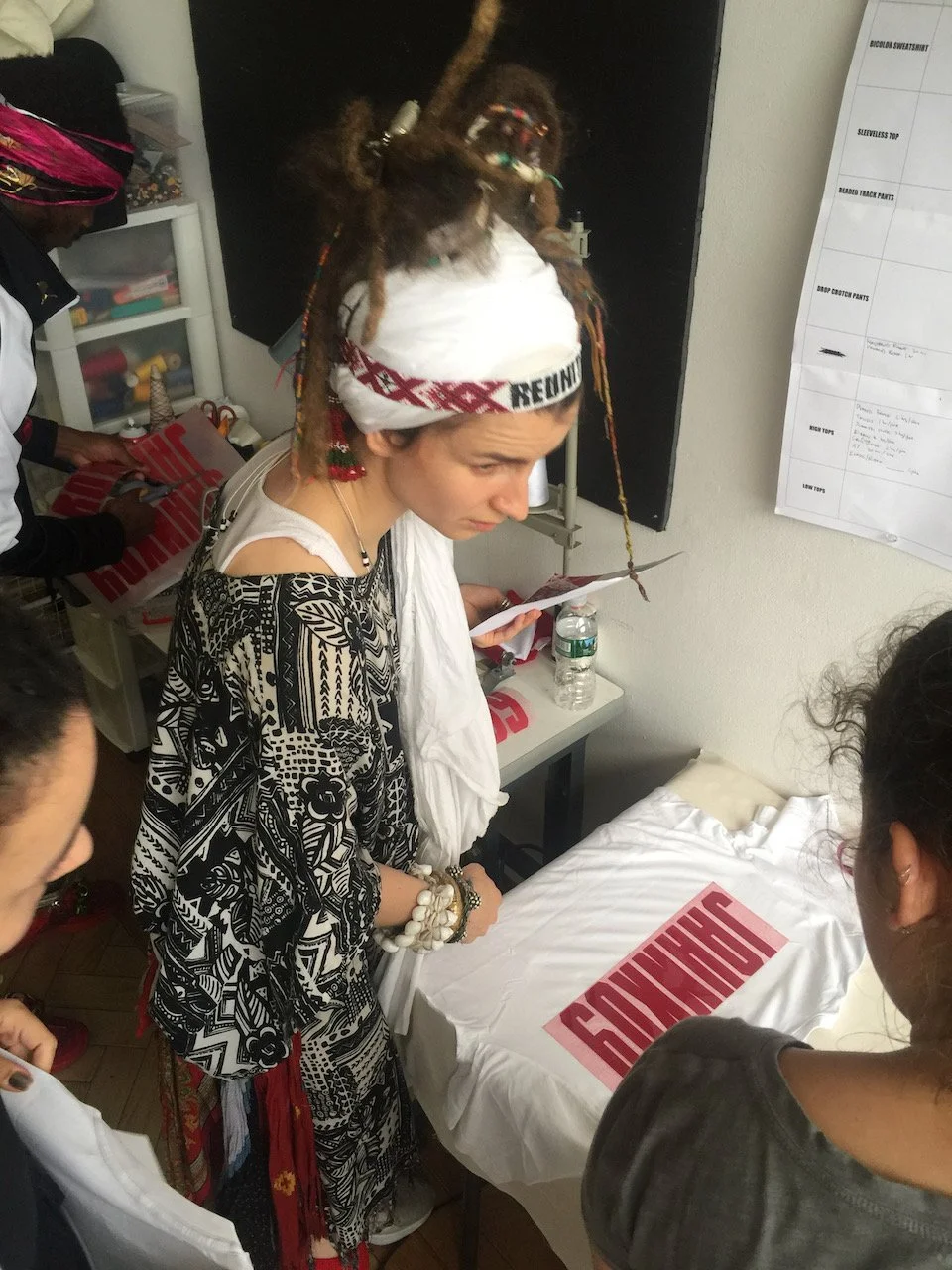 Young woman with dreadlocks and a beaded headband standing in a room, examining a white T-shirt with red text, surrounded by others, some holding additional T-shirts in a sewing or crafting space.