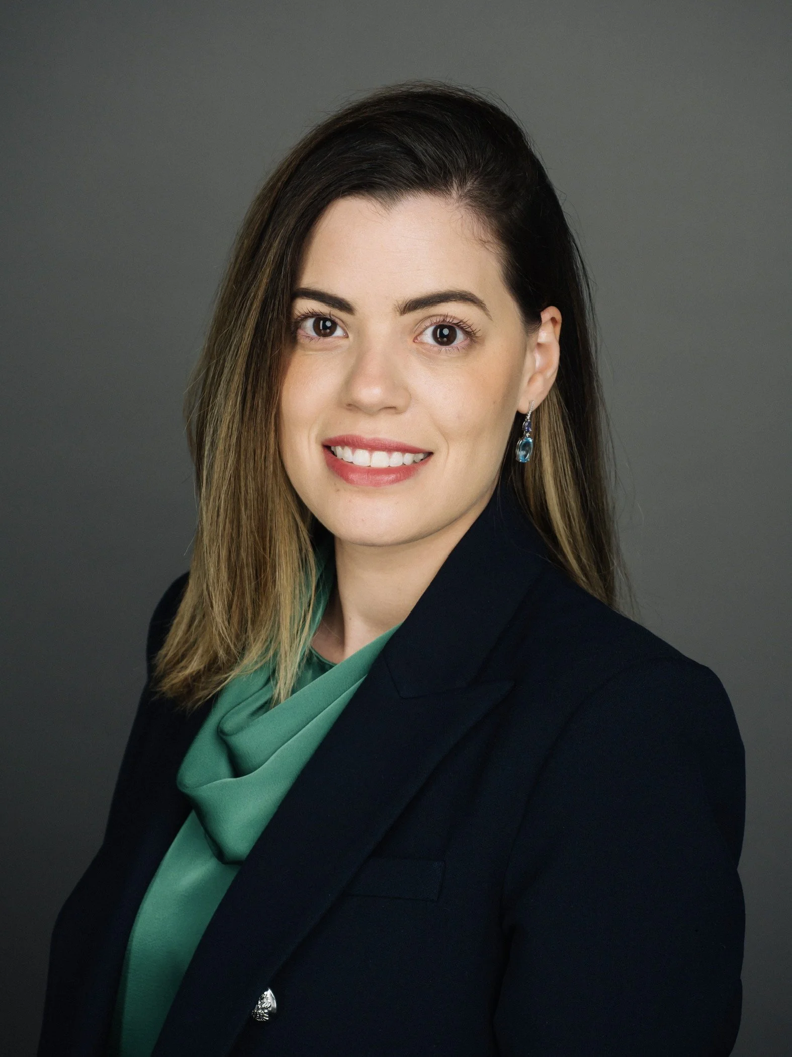 Professional woman with dark brown hair, wearing a black blazer, green blouse, and blue earrings, smiling against a gray background.