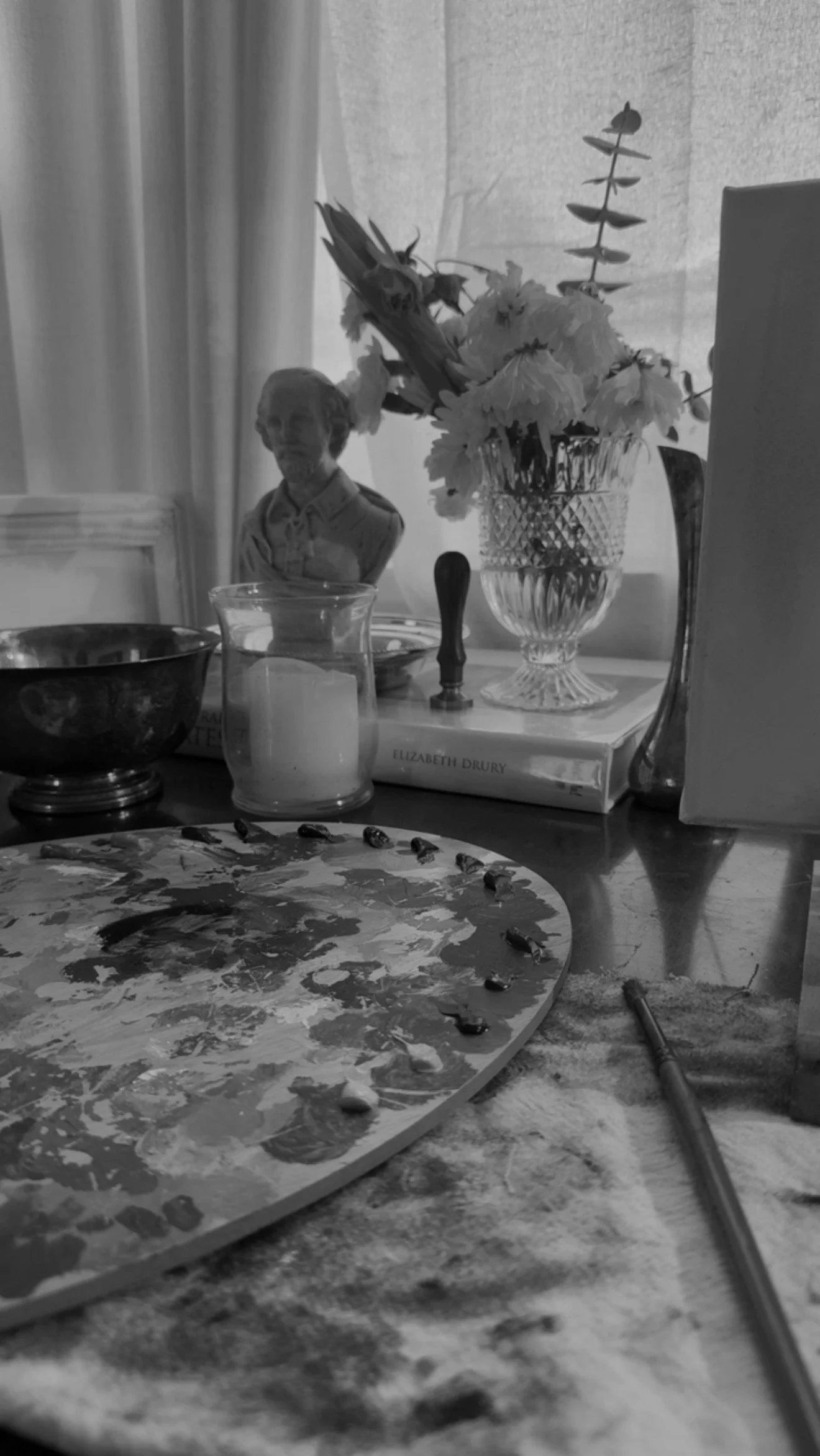 Black and white photo of a table with a paint palette, paintbrush, flowers in a glass vase, a sculpture of a child's bust, and a candle in a glass holder.