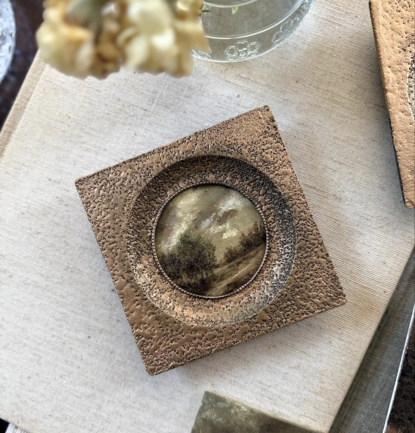 A square decorative frame with a textured, rough surface surrounding a circular mirror reflecting a landscape scene with trees and cloudy sky.