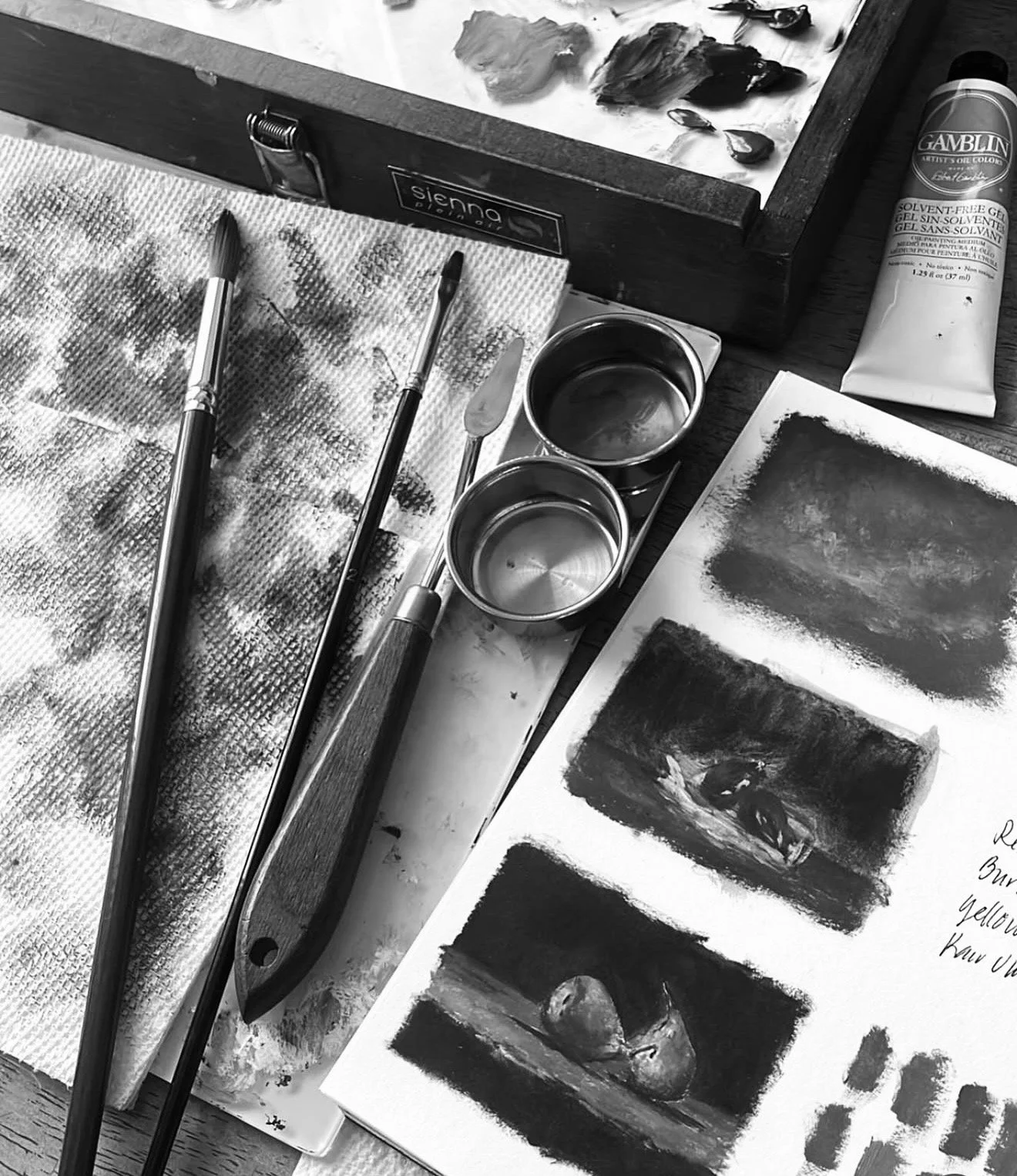 Black and white photograph of an artist's workspace with paintbrushes, a palette, small containers, a watercolor paper, and a tube of Gamblin solvent-free gel paint.