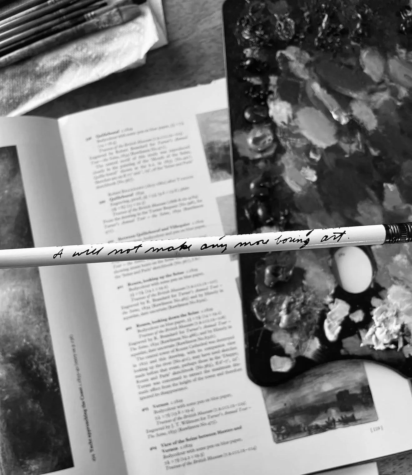 A black and white photo of an open book, a pencil with a handwritten message, a cutting board with pebbles, a flower, an egg, and some decorative items on a wooden table.