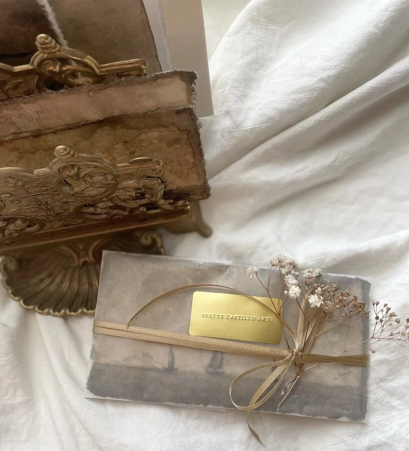 A vintage jewelry box with gold accents, a marble box with dried flowers and a gold label reading 'Ivette Castillo Art' on a white fabric surface.