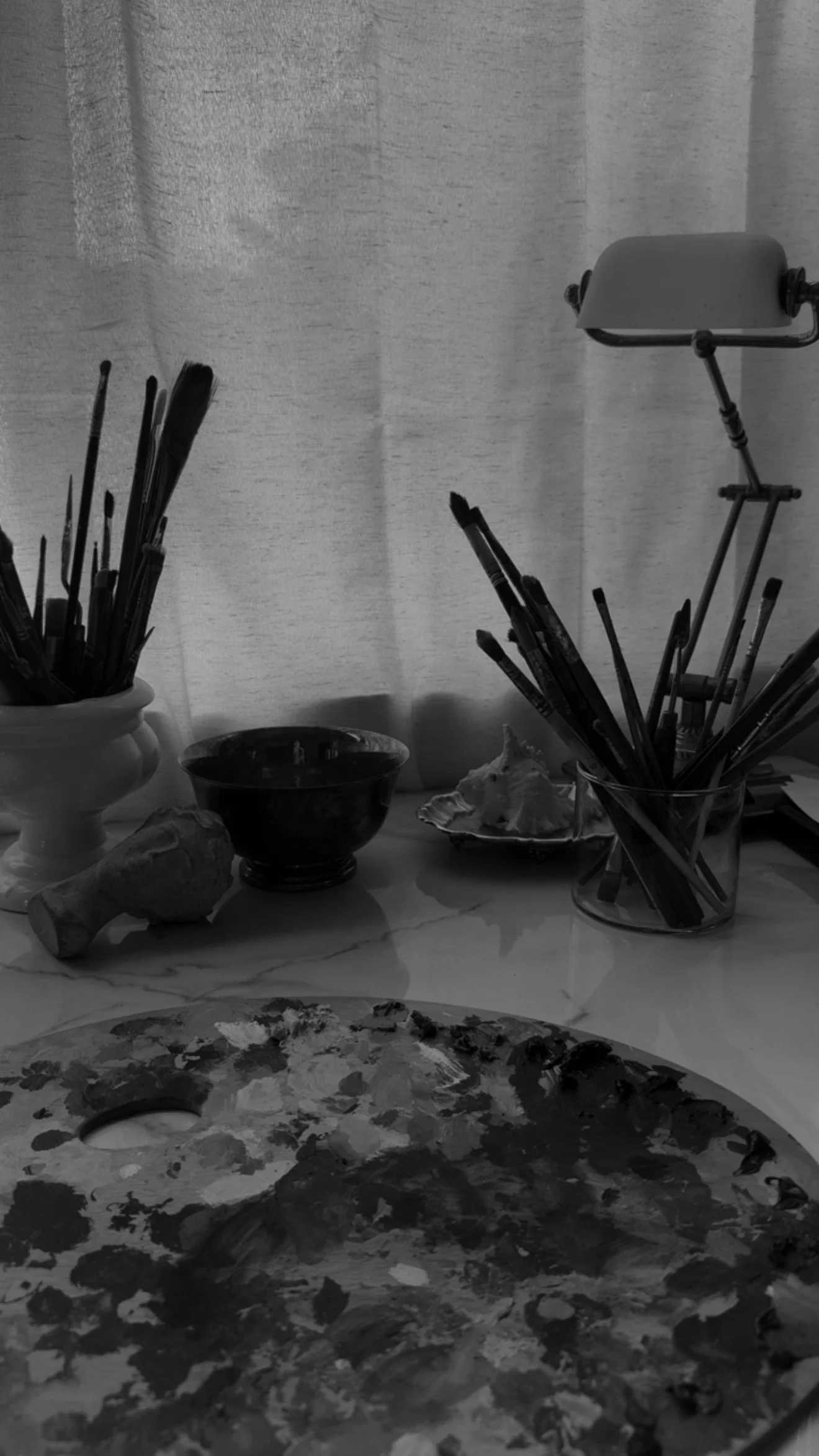 A table with art supplies including paintbrushes in containers, a palette with paint, a small bowl, and a tissue or sponge, in front of a window with curtains.