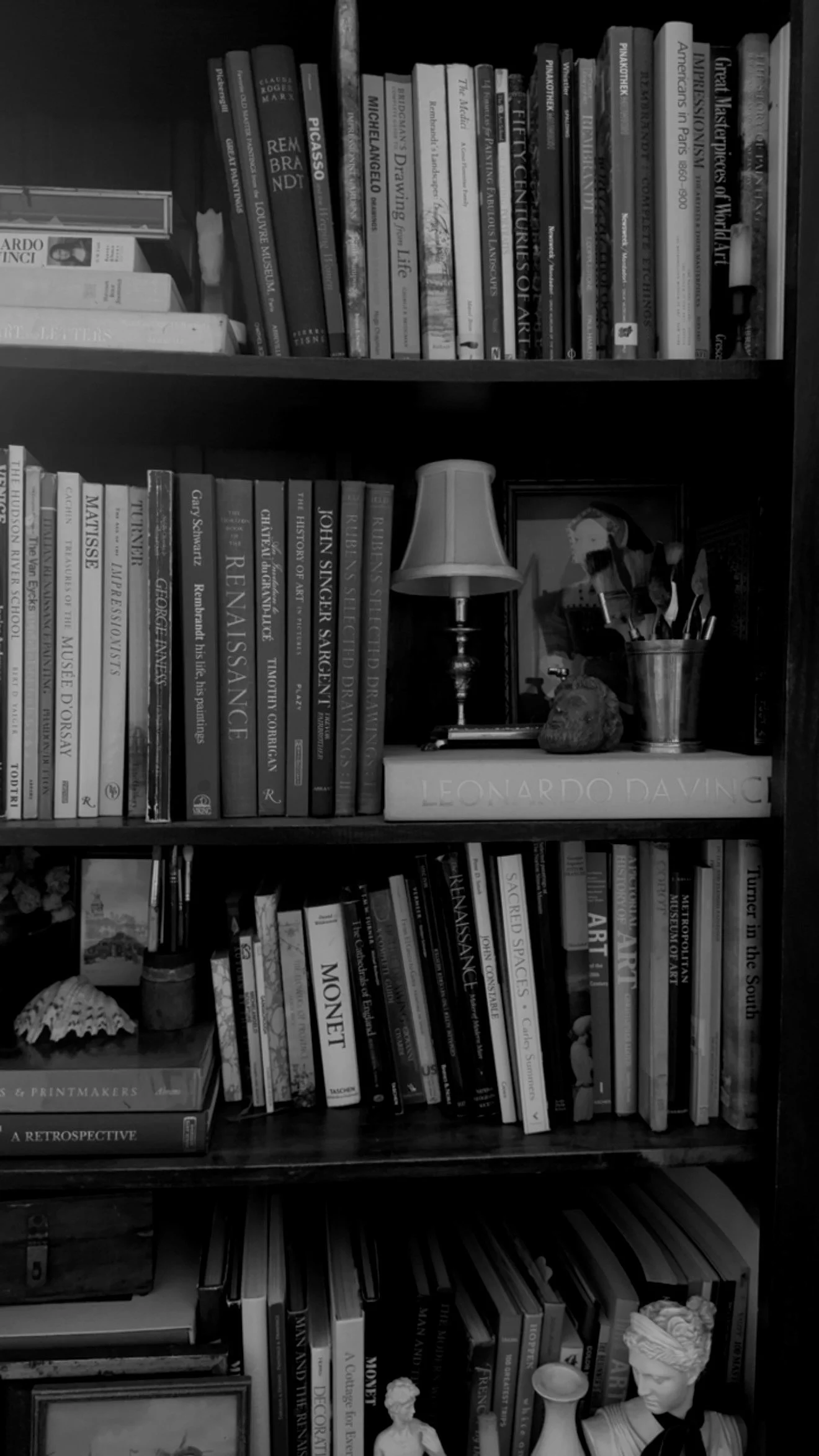 A bookshelf filled with art books, photography books, and art sculptures, including a small lamp and a potted plant on one shelf.