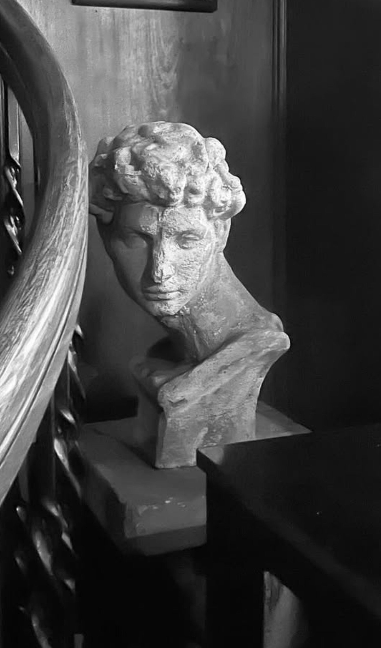 A grayscale photo of a bust sculpture of a woman with curly hair, placed on a pedestal between a wooden table and a dark wall.