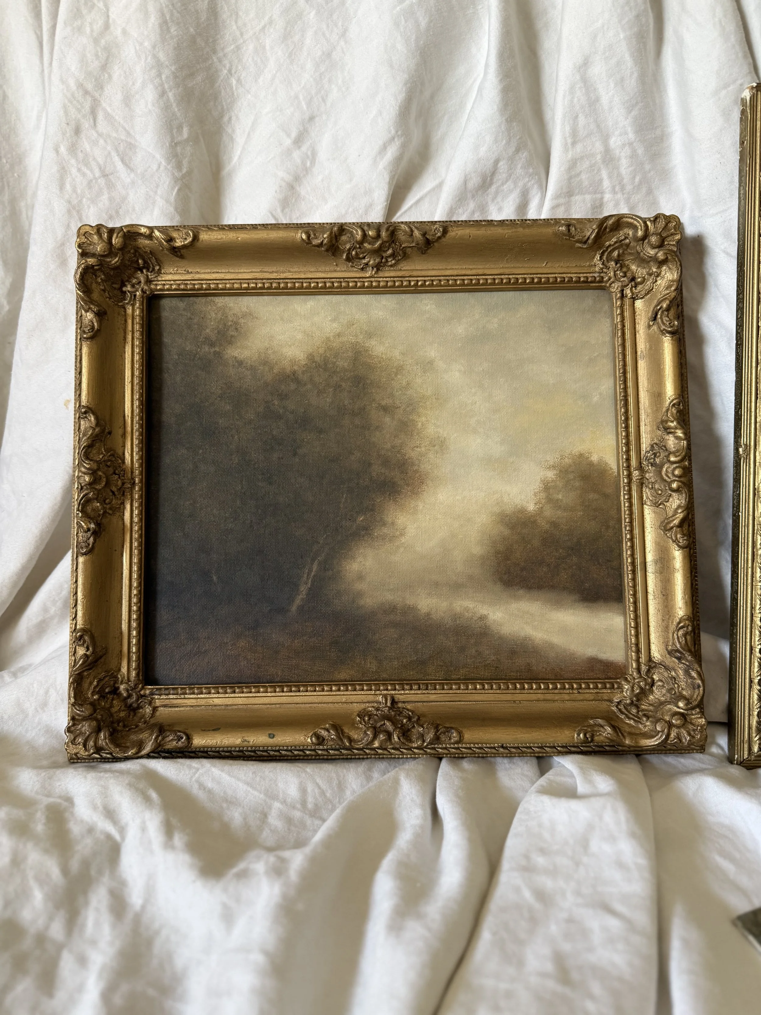 A framed landscape painting of trees and a river, in a gold ornate frame, resting on white fabric.
